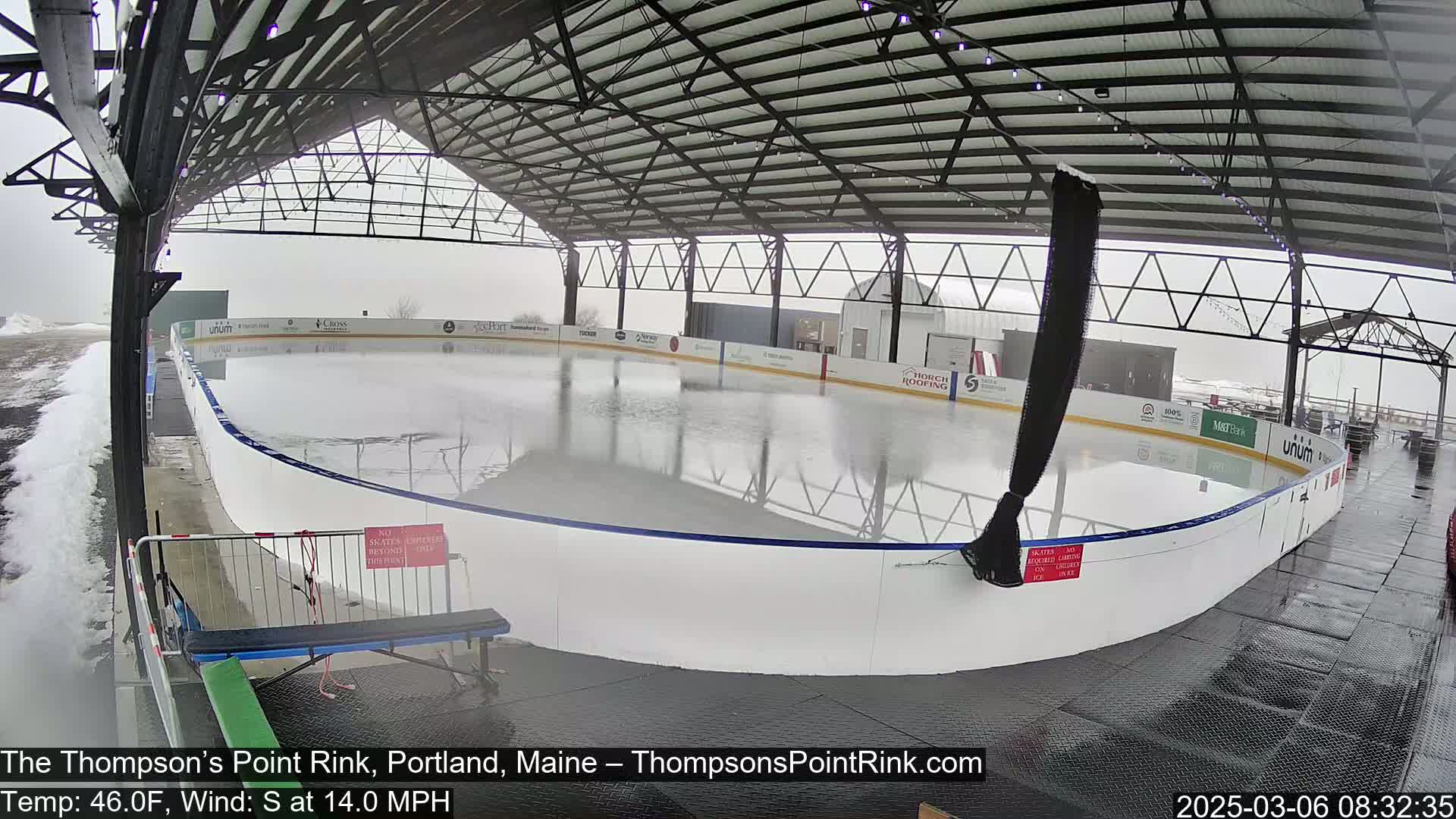 An outdoor ice rink, partially covered by a metal structure, is shown in rainy conditions.