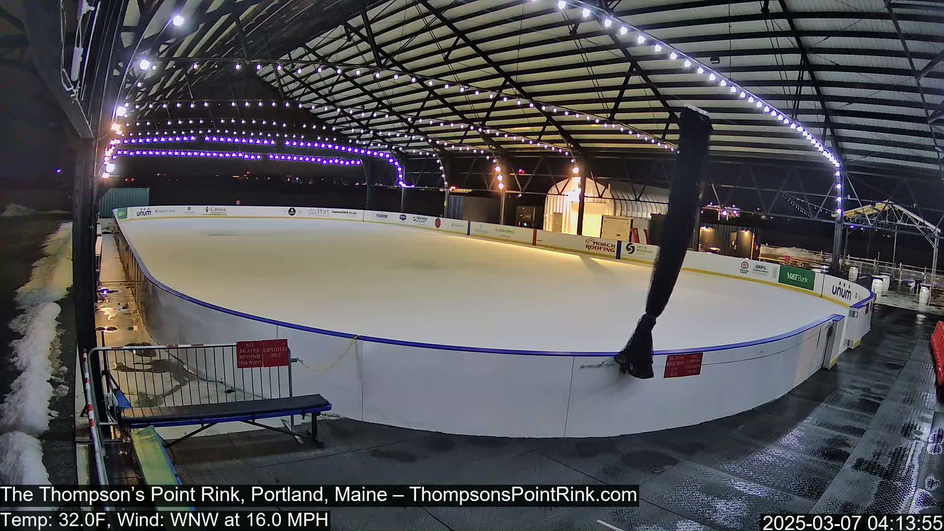 An outdoor ice rink, illuminated by overhead lights, is mostly empty under a nighttime sky with a temperature of 32°F and a 16 mph wind from the west-northwest.