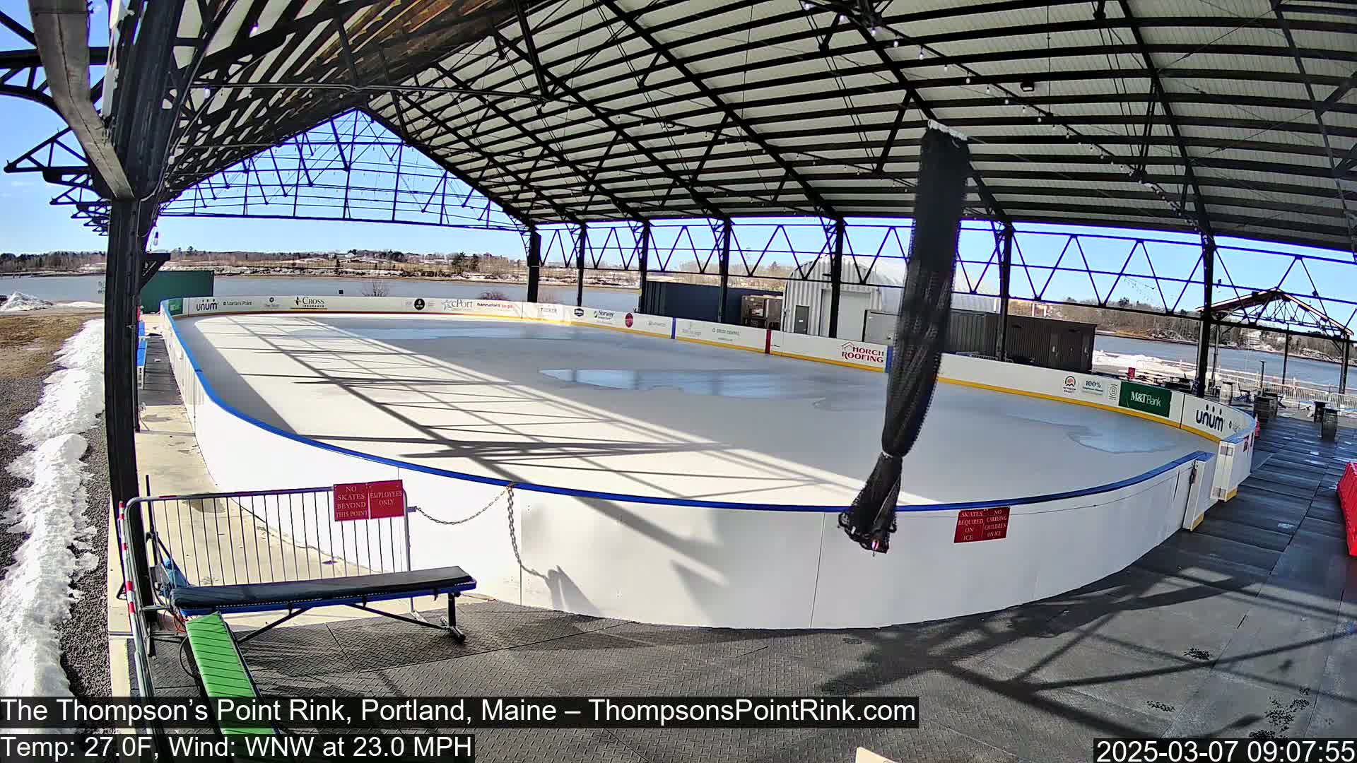 An outdoor ice rink, under a metal roof structure, on a sunny day with a strong wind.
