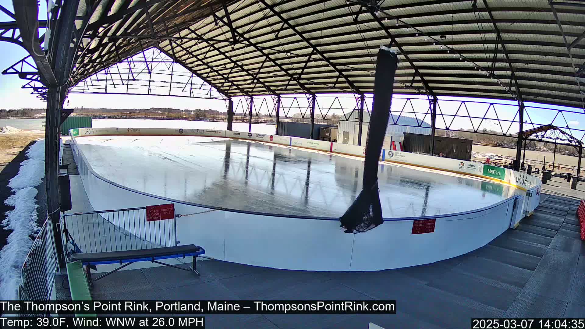 An outdoor ice rink, partially covered by a metal structure, is visible on a sunny day with a 26 mph wind.