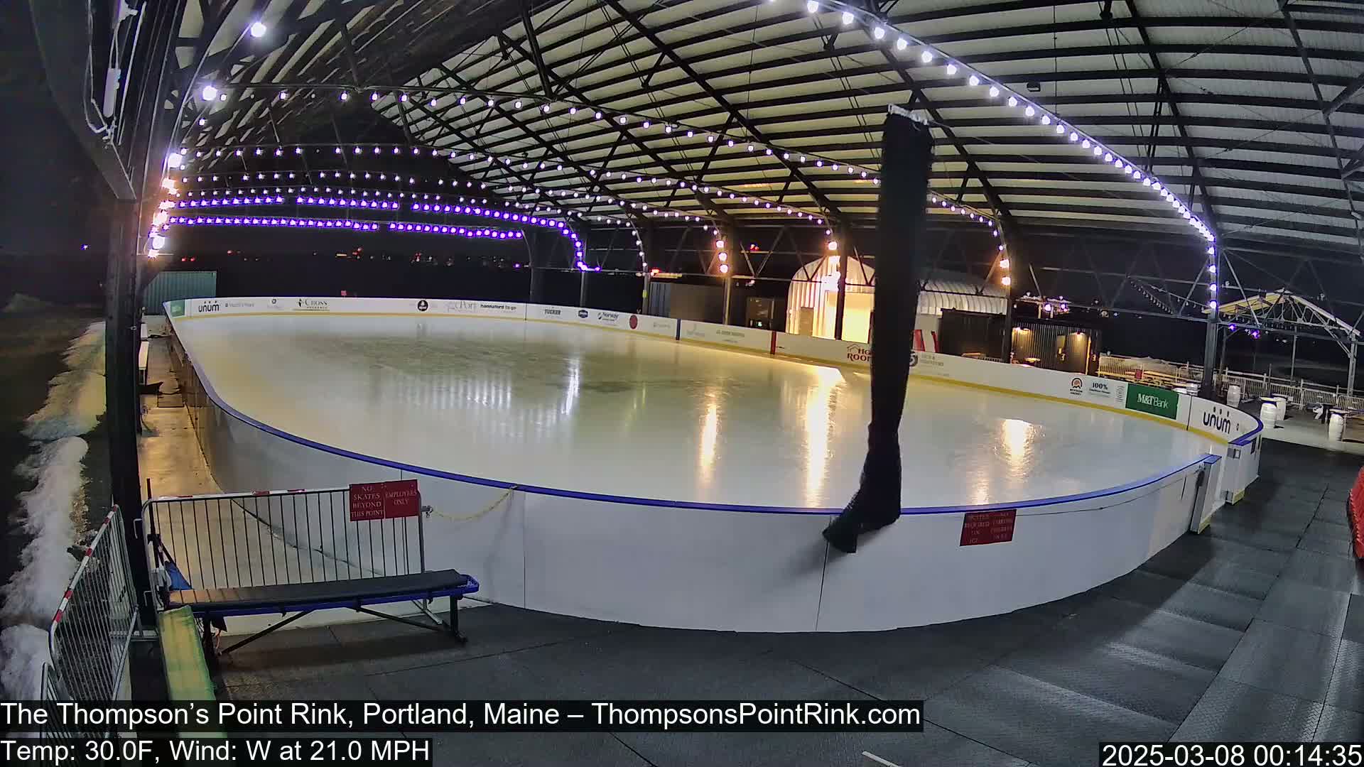 An outdoor ice rink, illuminated at night under a covered structure, is surrounded by choppy water.
