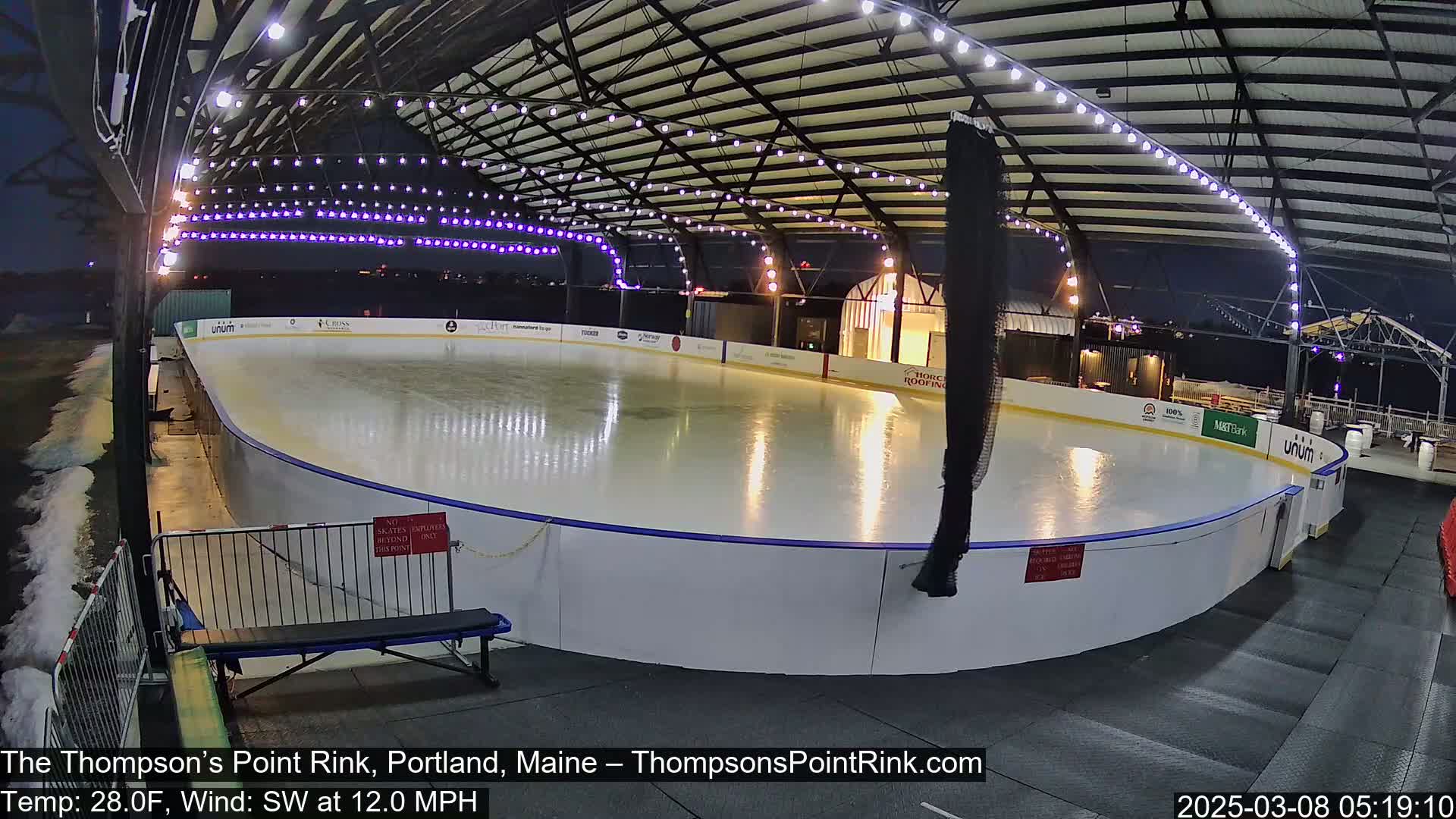 An outdoor ice rink, illuminated at night under a covered structure, with a southwest wind blowing at 12 mph and a temperature of 28 degrees Fahrenheit.