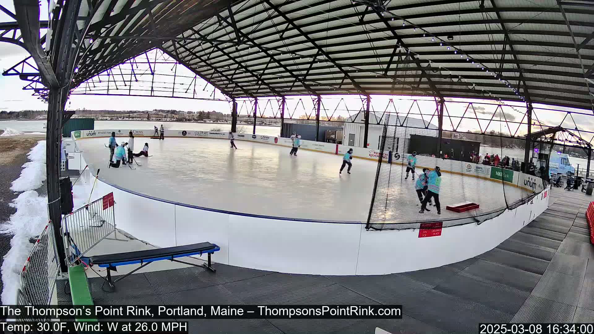 An outdoor ice rink, covered by a metal structure, shows several people playing hockey in windy, 30°F weather.
