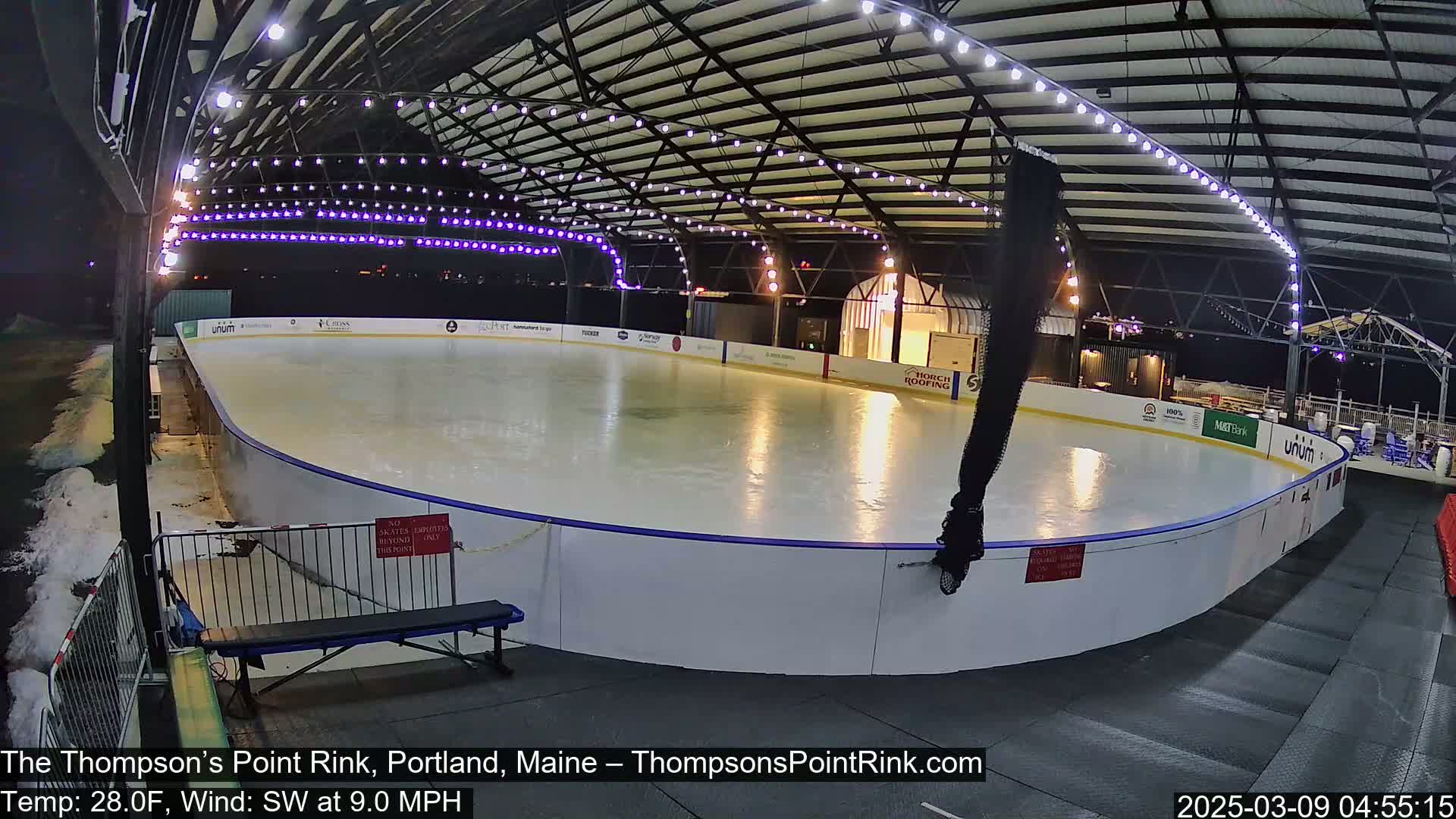 An outdoor ice rink, illuminated at night under a metal structure with string lights, is mostly empty with a person visible near the rink's edge under a southwest wind of 9 mph and a temperature of 28°F.