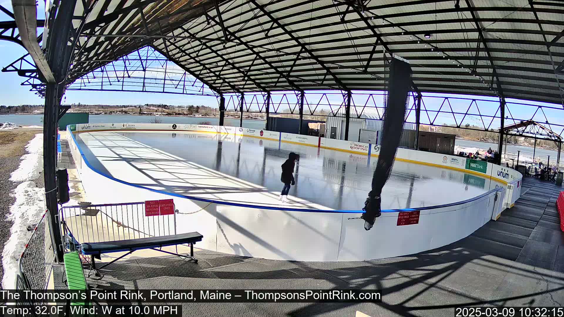 A person ice skates on an outdoor rink under a metal-framed shelter on a sunny day with a 10 mph westerly wind and a temperature of 32°F.