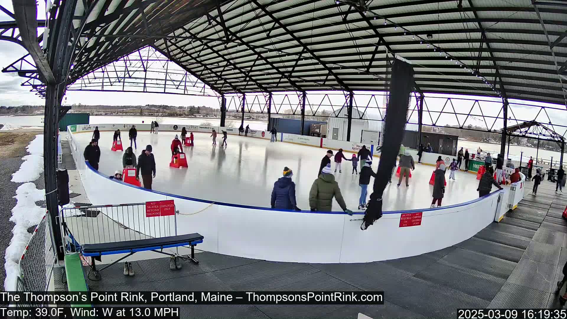 Many people ice skate on an outdoor rink under a covered structure on a partly cloudy day with a westerly wind.