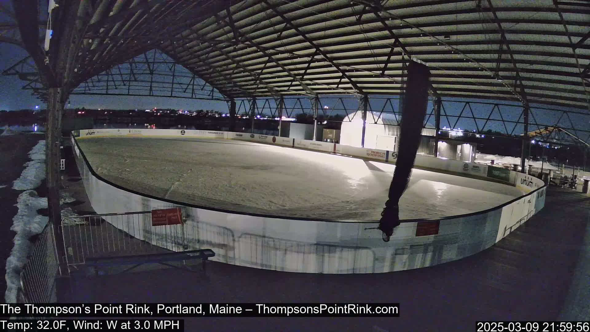 An outdoor ice rink, mostly empty under a partially covered structure at night, with light snow or ice accumulation visible around the rink and a slight wind.