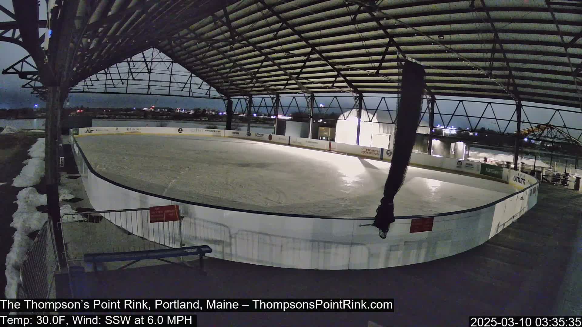 An outdoor ice rink, partially covered by a metal structure, is mostly empty under a dark, nighttime sky with a temperature of 30 degrees Fahrenheit and a south-southwesterly wind.
