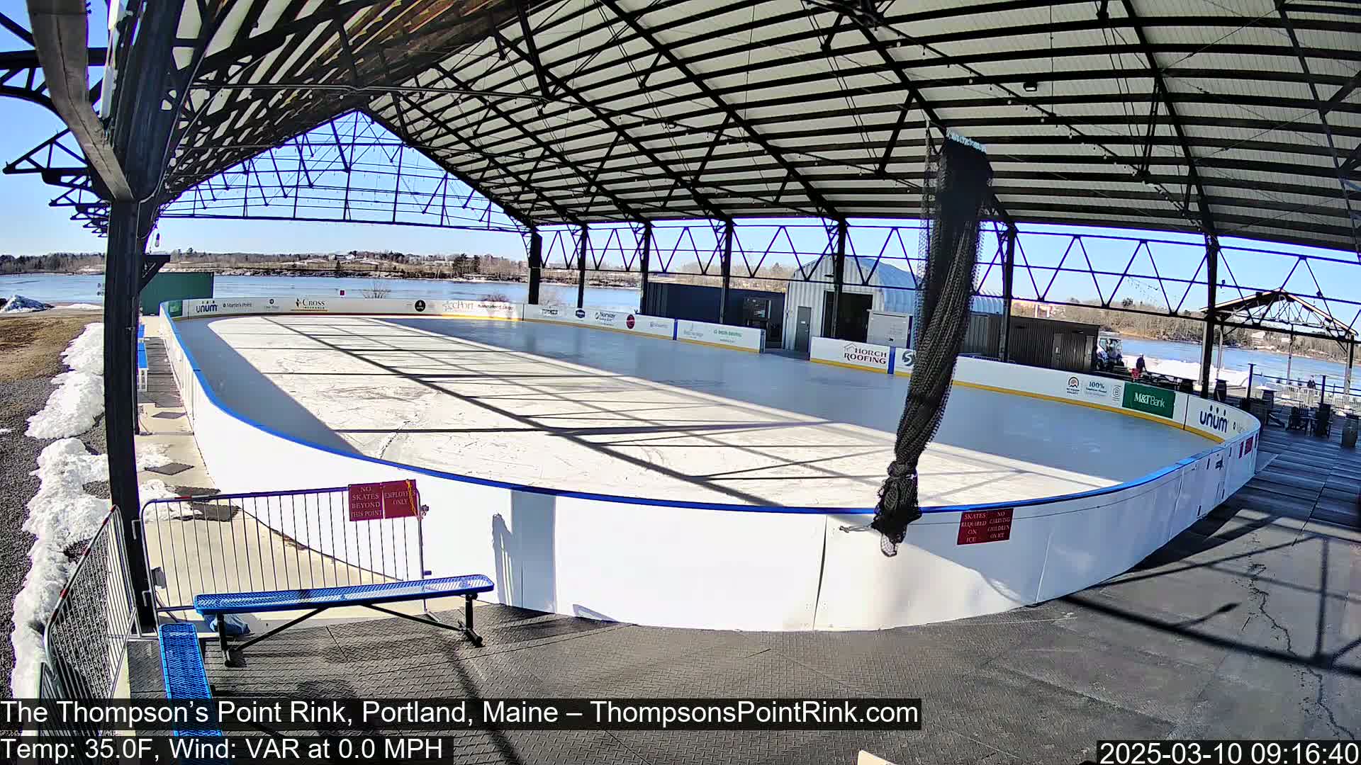 An outdoor ice rink, under a metal framework, on a sunny day with some snow on the ground.