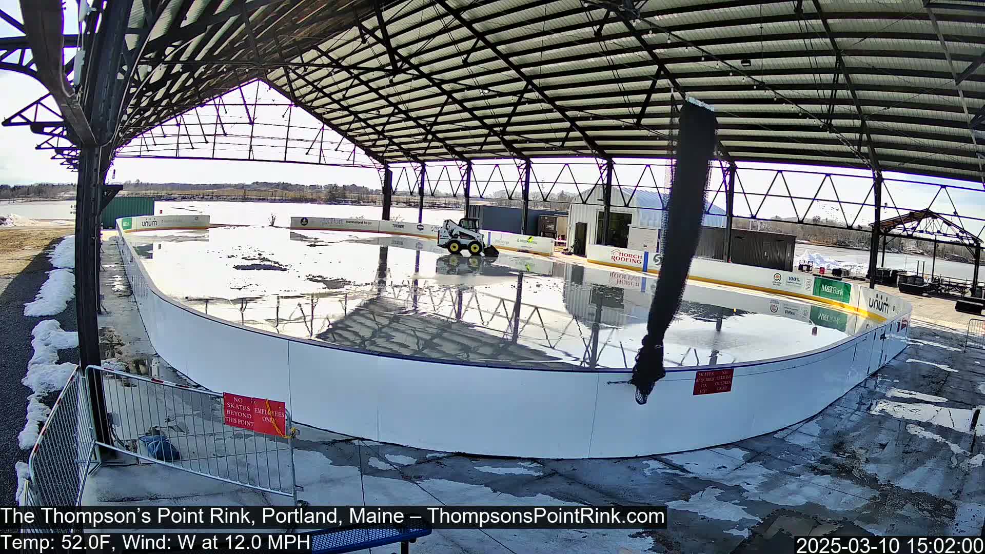 A wet outdoor ice rink under a metal roof is being serviced by a small piece of machinery on a sunny day with a 12 mph west wind.