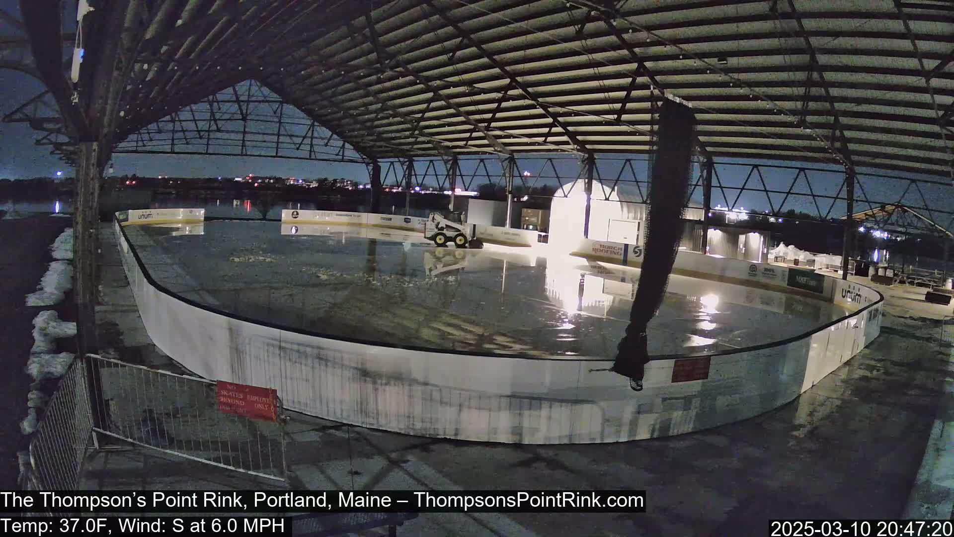 An outdoor ice rink under a metal structure at night is being lightly rained on, with a small machine on the ice and some snow visible nearby.