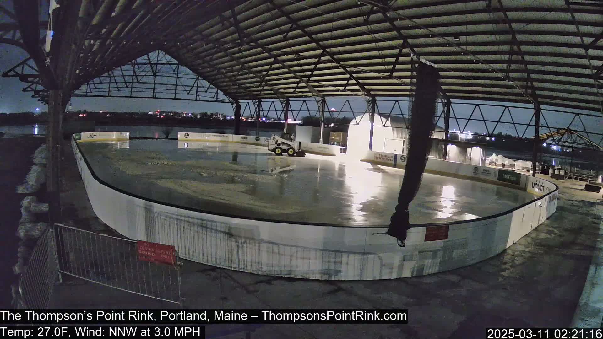 A partially flooded outdoor ice rink sits under a metal structure at night, with a small piece of machinery on the ice.