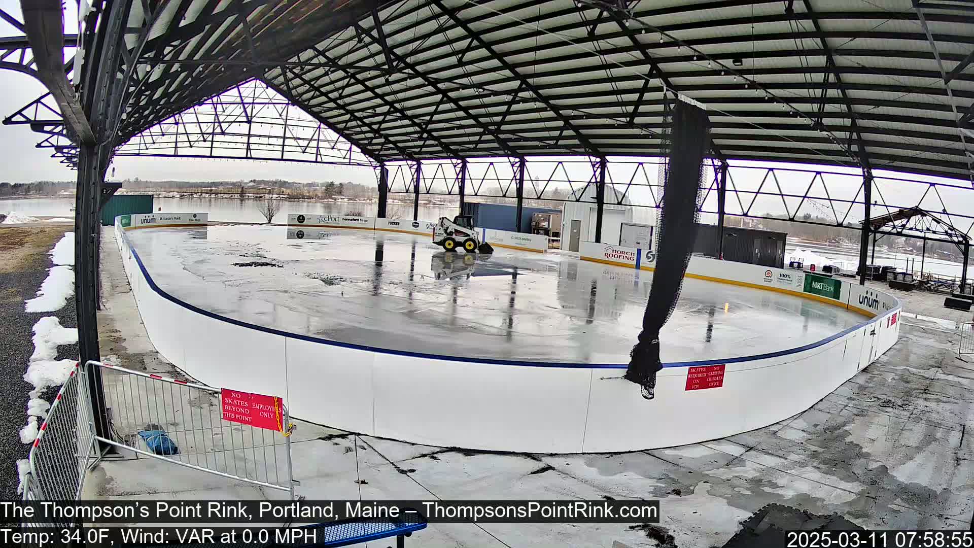 An outdoor rink under a metal structure is being cleaned by a small machine; there is snow on the ground and the weather is 34°F with variable wind at 0.0 mph.