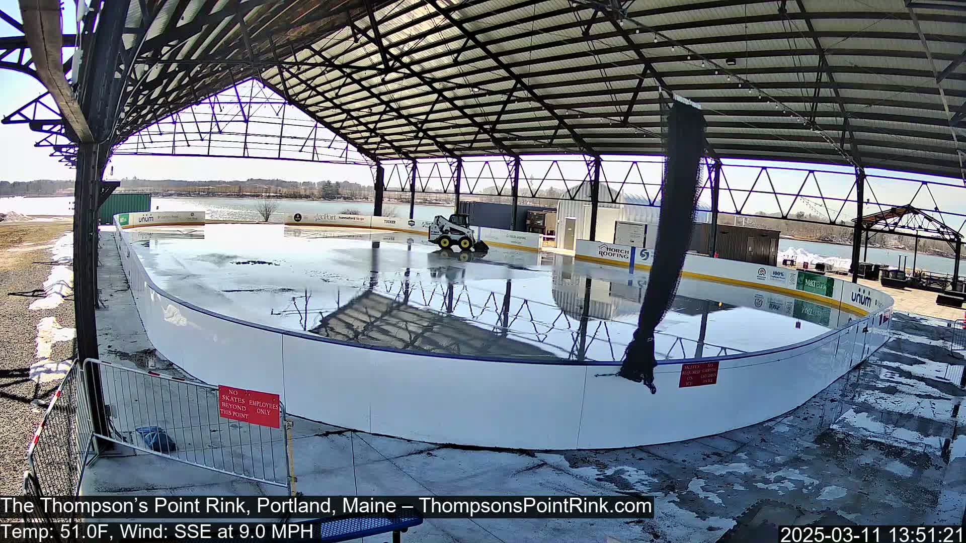 A wet outdoor ice rink under a metal roof is being serviced by a small piece of machinery on a sunny day with a 9 mph wind.