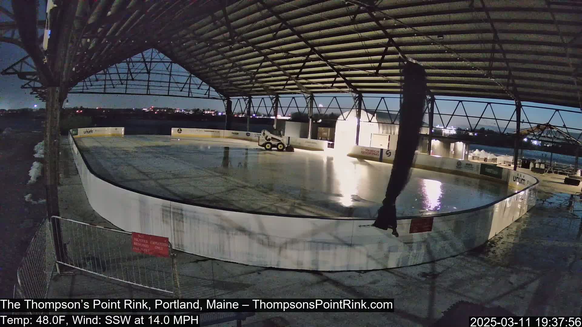 A wet outdoor ice rink is under a metal roof structure at night with a small piece of machinery on the ice and a 14 mph wind from the south-southwest.