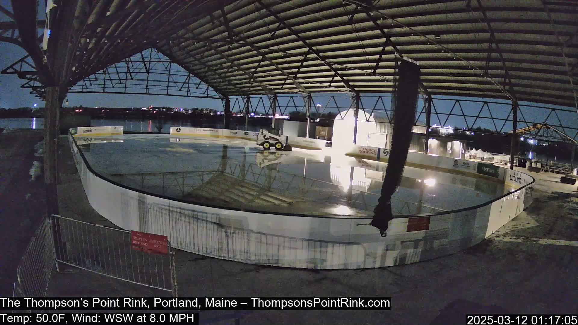 An outdoor ice rink under a metal structure at night, with a small vehicle parked on the ice and a calm, 50°F temperature, and a light WSW wind.