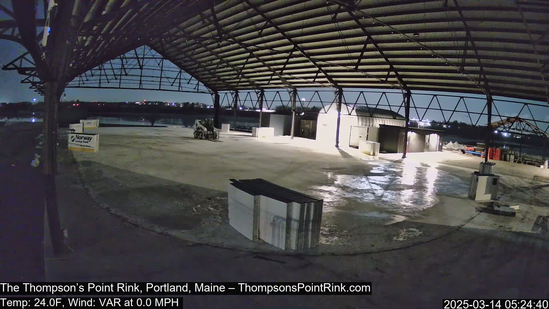 Under a dark night sky, a large empty metal-framed structure sits on a wet concrete surface, with some construction materials and equipment visible.