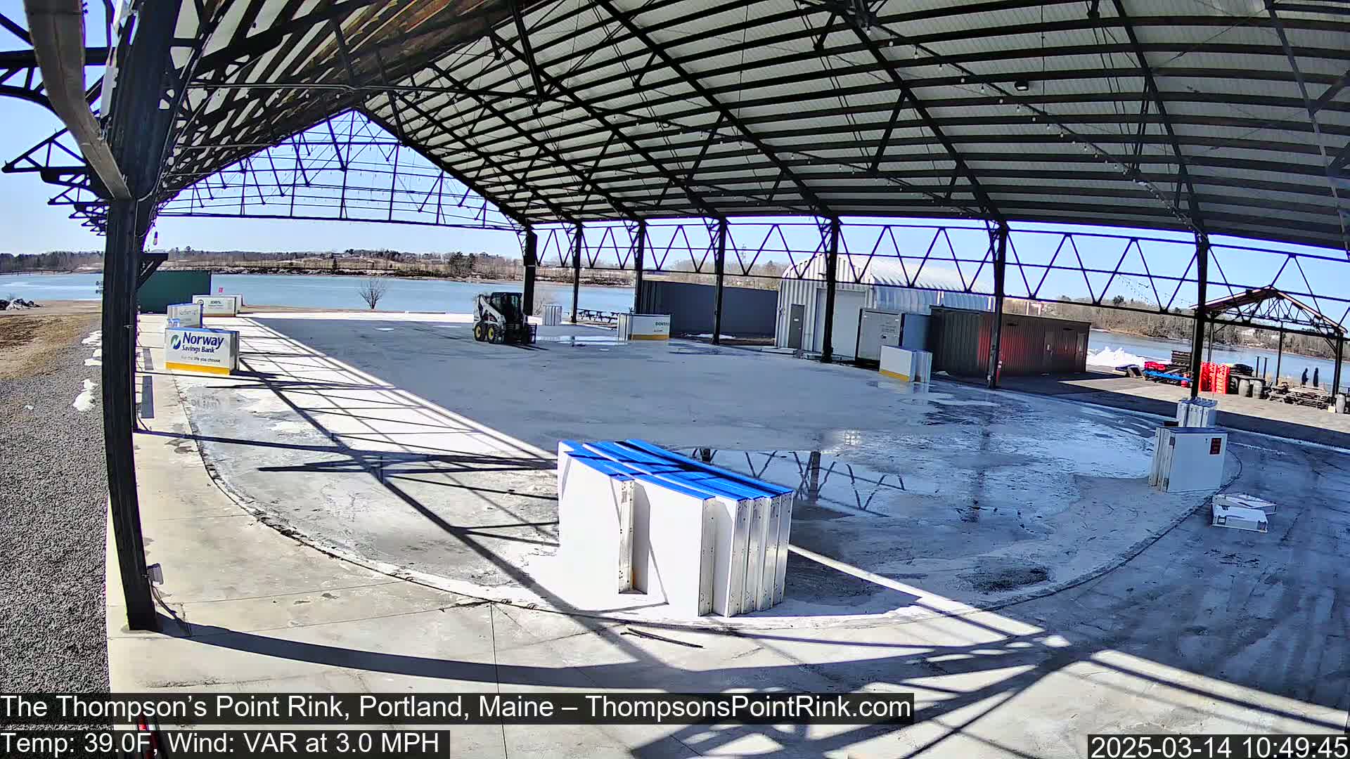 An empty, open-air rink under a metal roof on a sunny day with a body of water visible in the background.