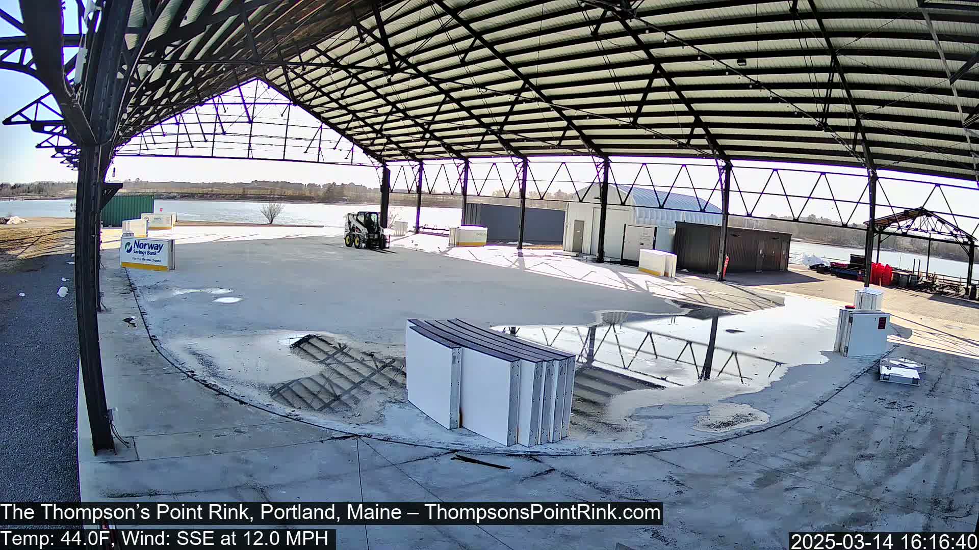 A large open-air structure with a metal roof and concrete floor shows a partly sunny day with a SSE wind of 12 mph and a temperature of 44°F; there's a puddle on the ground, and several white panels are stacked in the foreground.