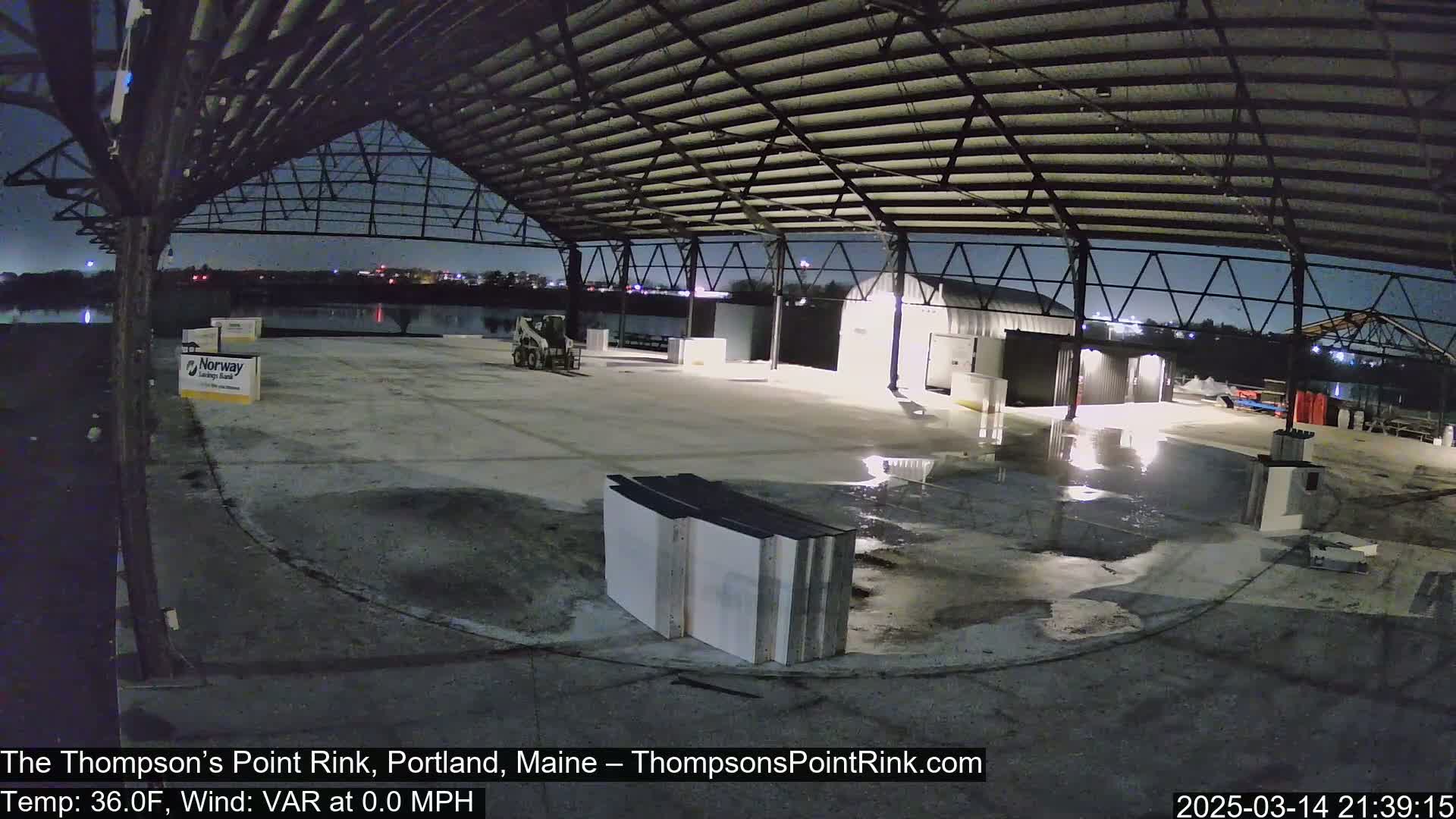 A large, empty, open-air structure with a metal roof is illuminated at night, showing some puddles on the concrete floor.