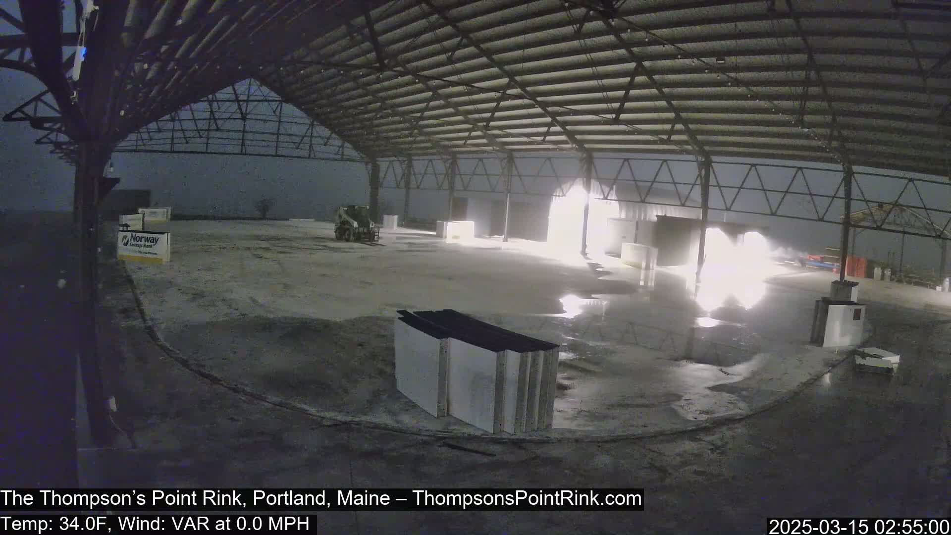 A nighttime view of an open-air structure's interior, showing a bright light source and wet ground under a dark, possibly snowy sky.