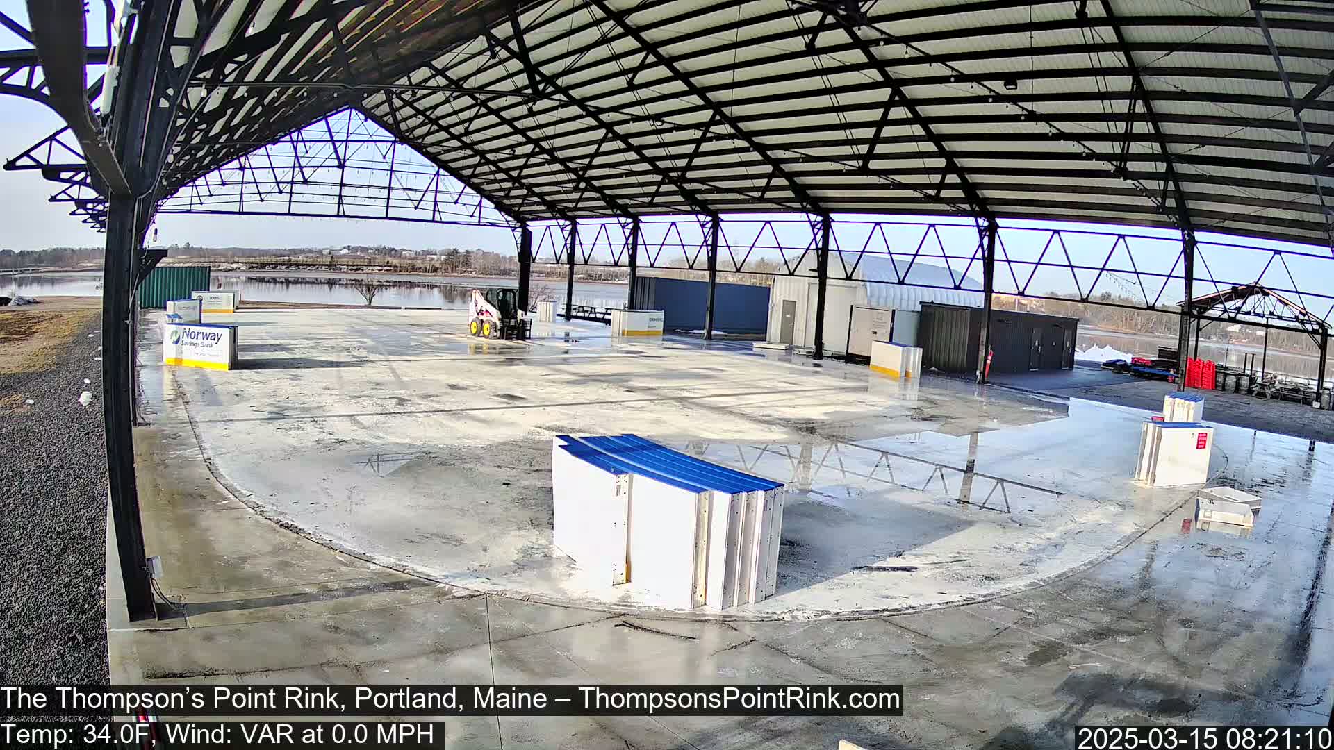 A wet, empty concrete rink under a large metal-framed structure on a sunny day.