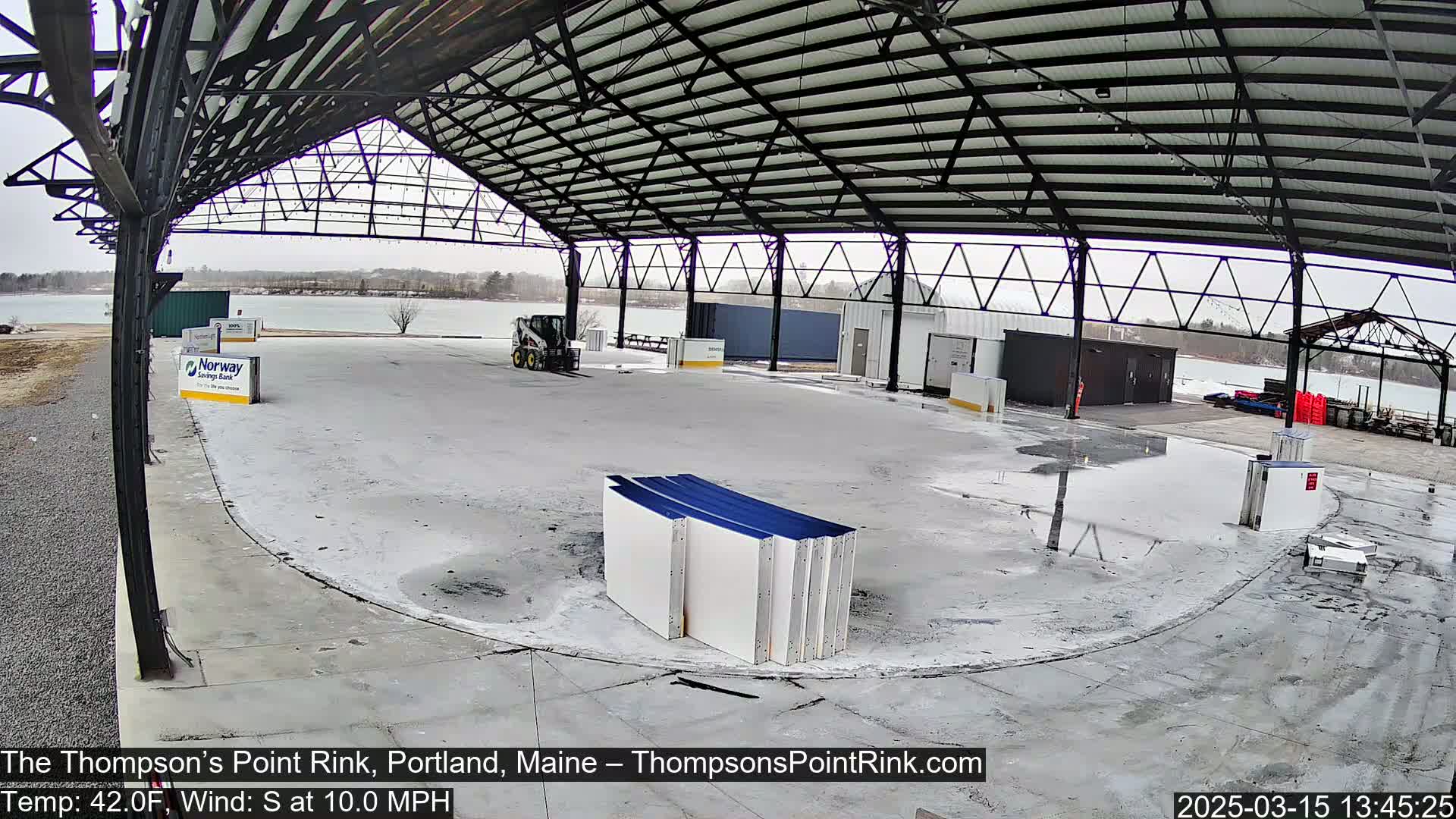 A large, open-air rink under a metal roof is partially covered in standing water, with a small skid steer and several stacked rink boards visible; it is a cloudy day.
