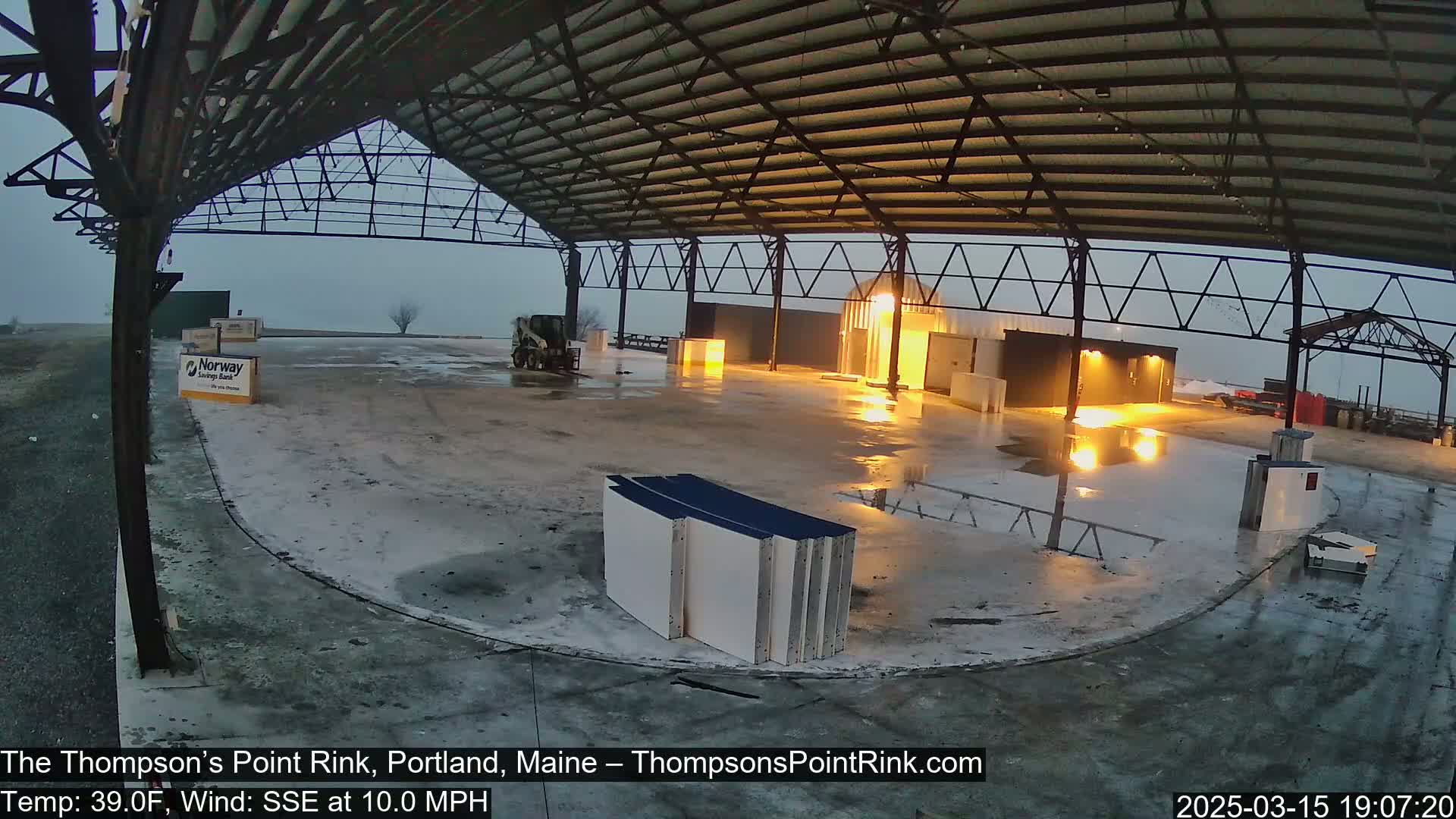 A wet, empty rink under a large metal roof in overcast conditions.