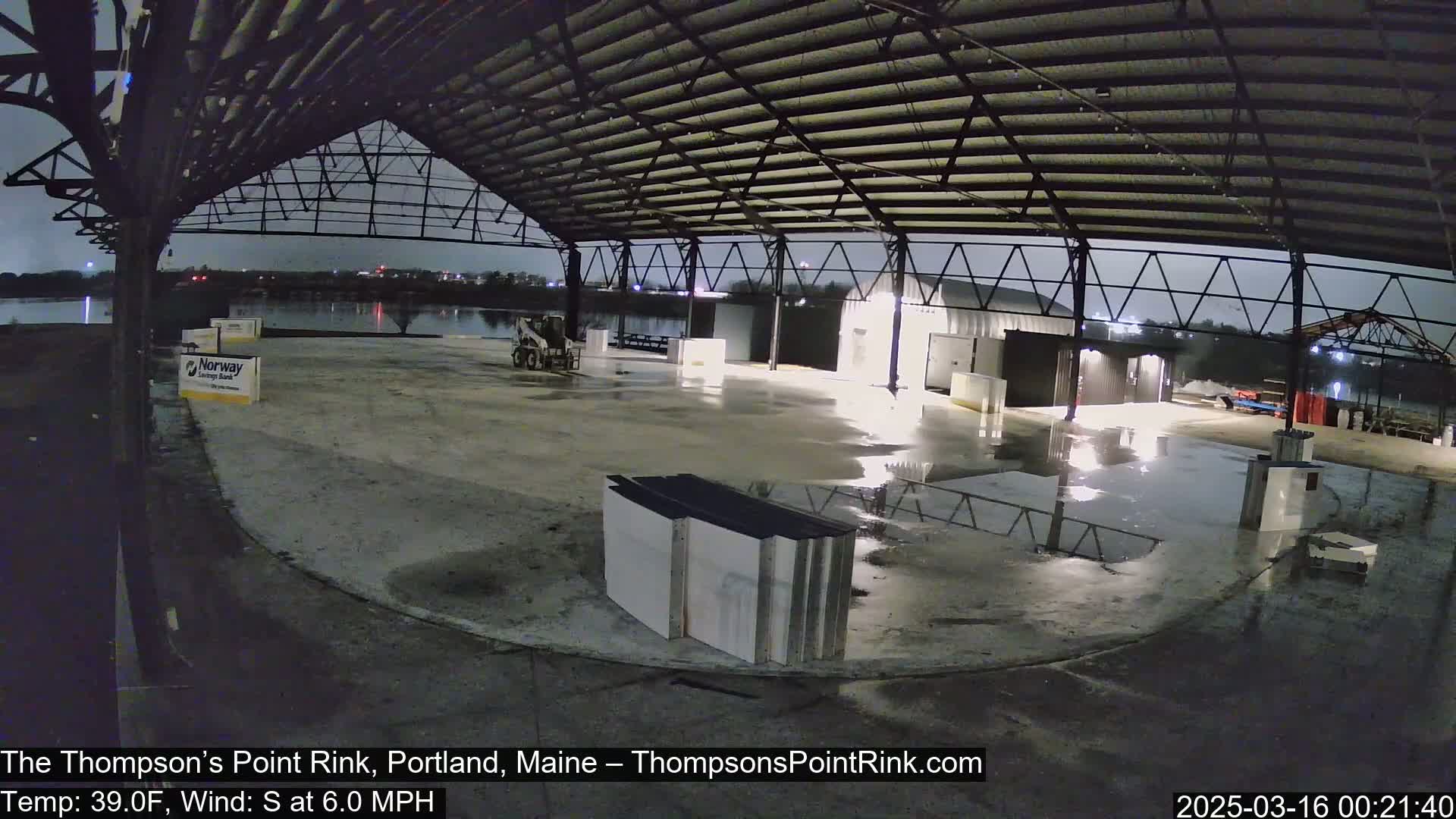A nighttime view of a large, open-air structure with a metal roof, wet ground, and some construction materials, under a dark sky with a south wind.