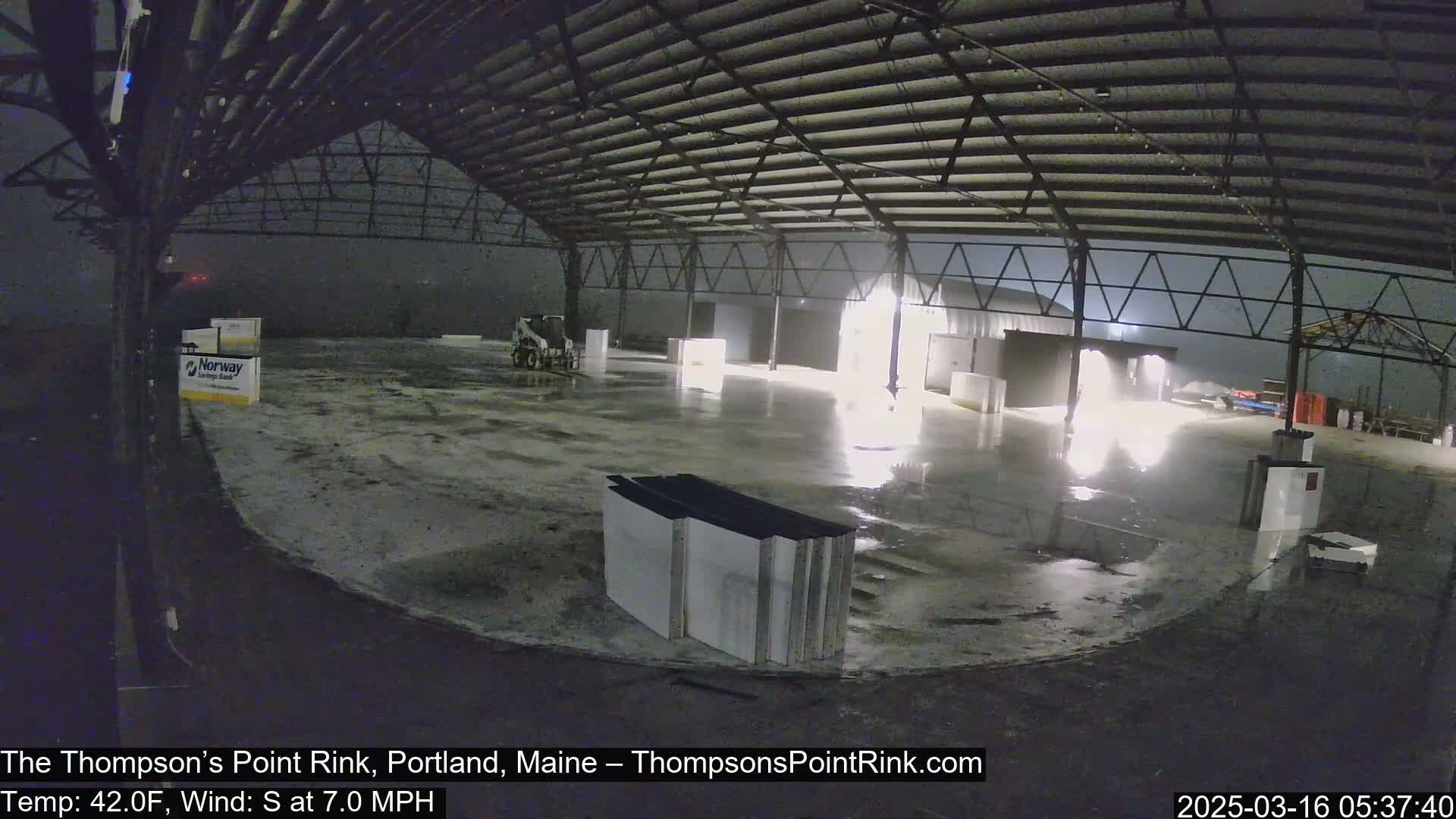 A large, empty indoor rink is seen at night during a light rain, with a small piece of construction equipment visible in the background.