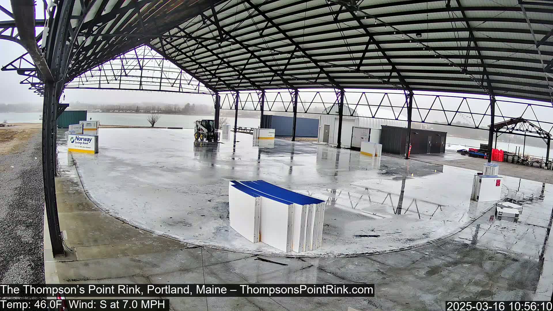 A wet, open-air rink under a metal framework, with a body of water visible in the background during overcast conditions.