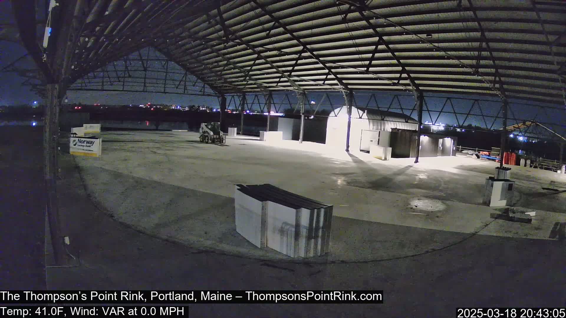An empty, open-air rink under a metal framework at night, illuminated by lights, with calm weather conditions.