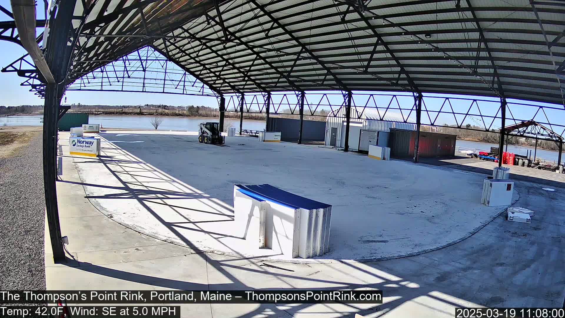 A large, empty, open-air rink under a metal roof on a sunny day with a calm, partly cloudy sky.