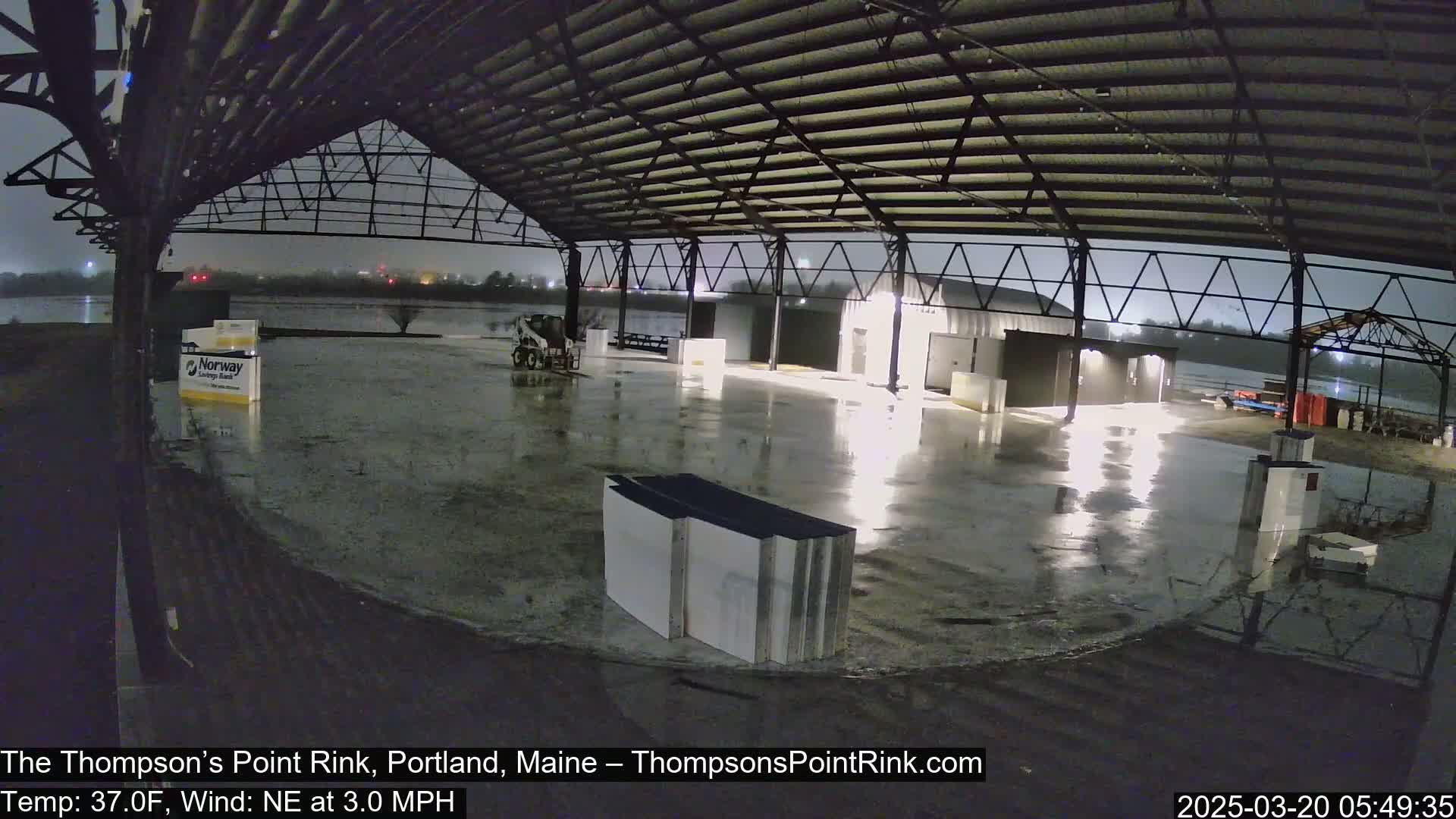 A large, empty, wet rink under a metal roof is illuminated at night by artificial lights during a light rain with a northeast wind.