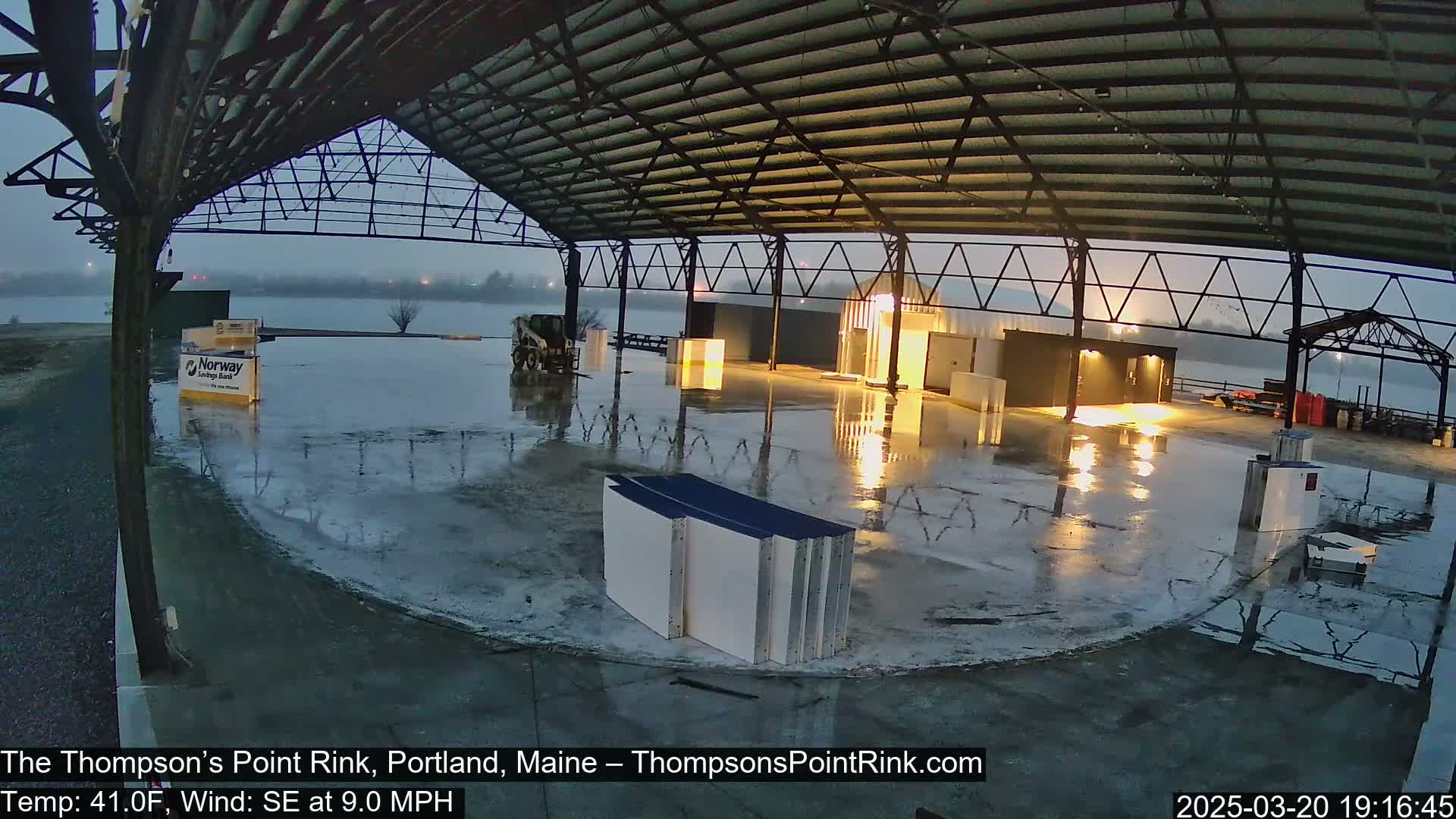 A wet, open-air rink under a metal framework is illuminated by artificial lights in the twilight.