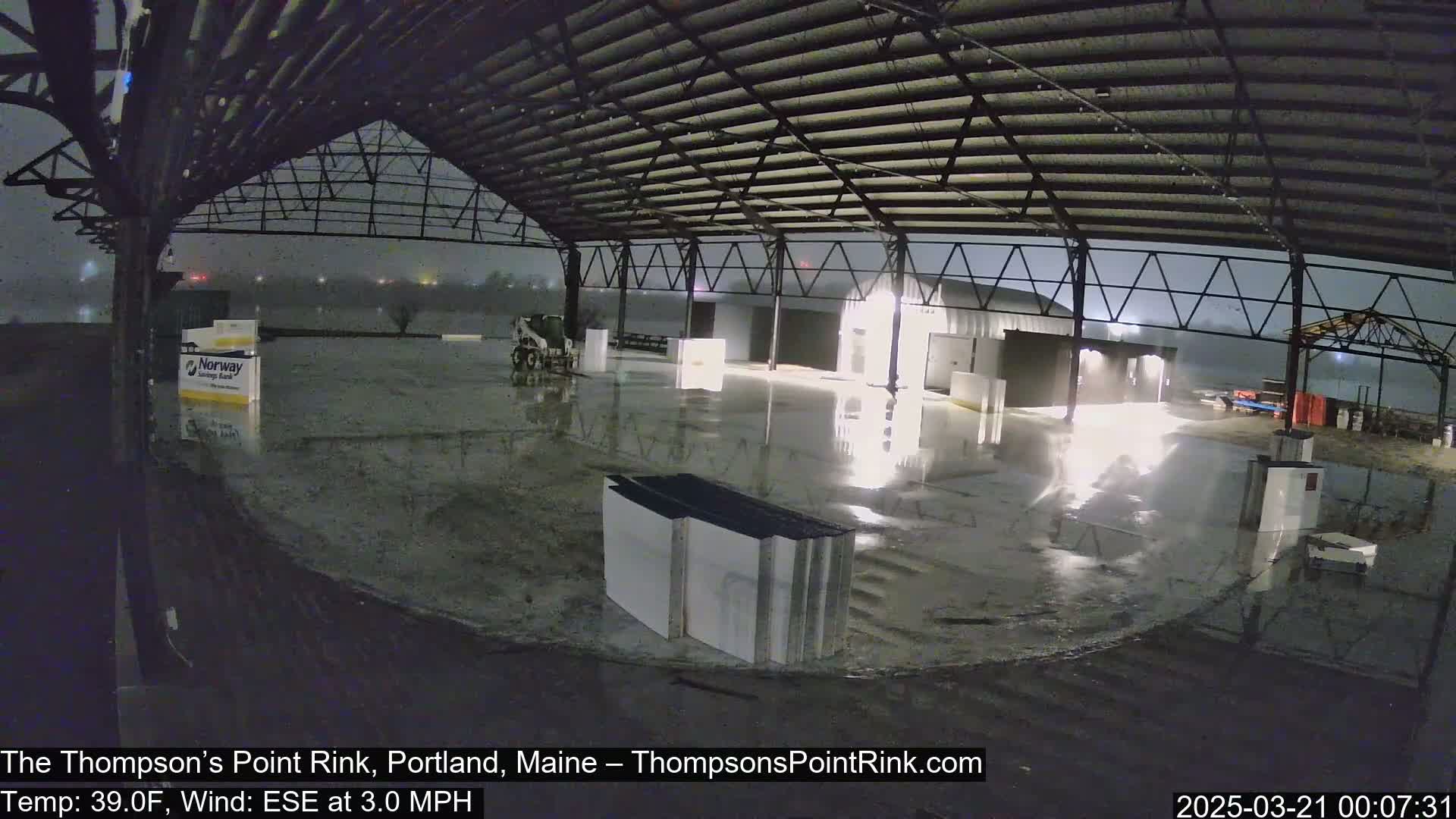 A wet, empty rink under a large metal roof is illuminated at night during a light rain.