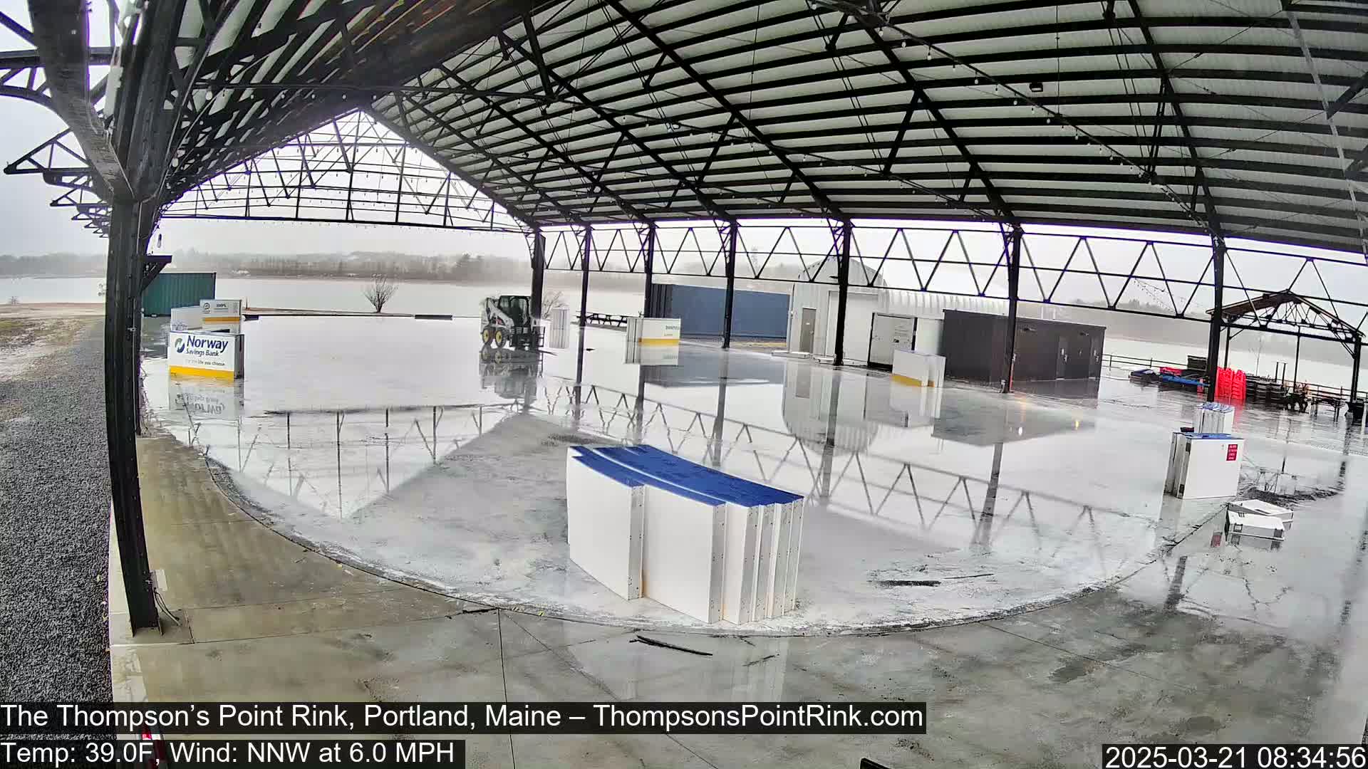A wet, empty rink inside a large metal structure on a cloudy day.