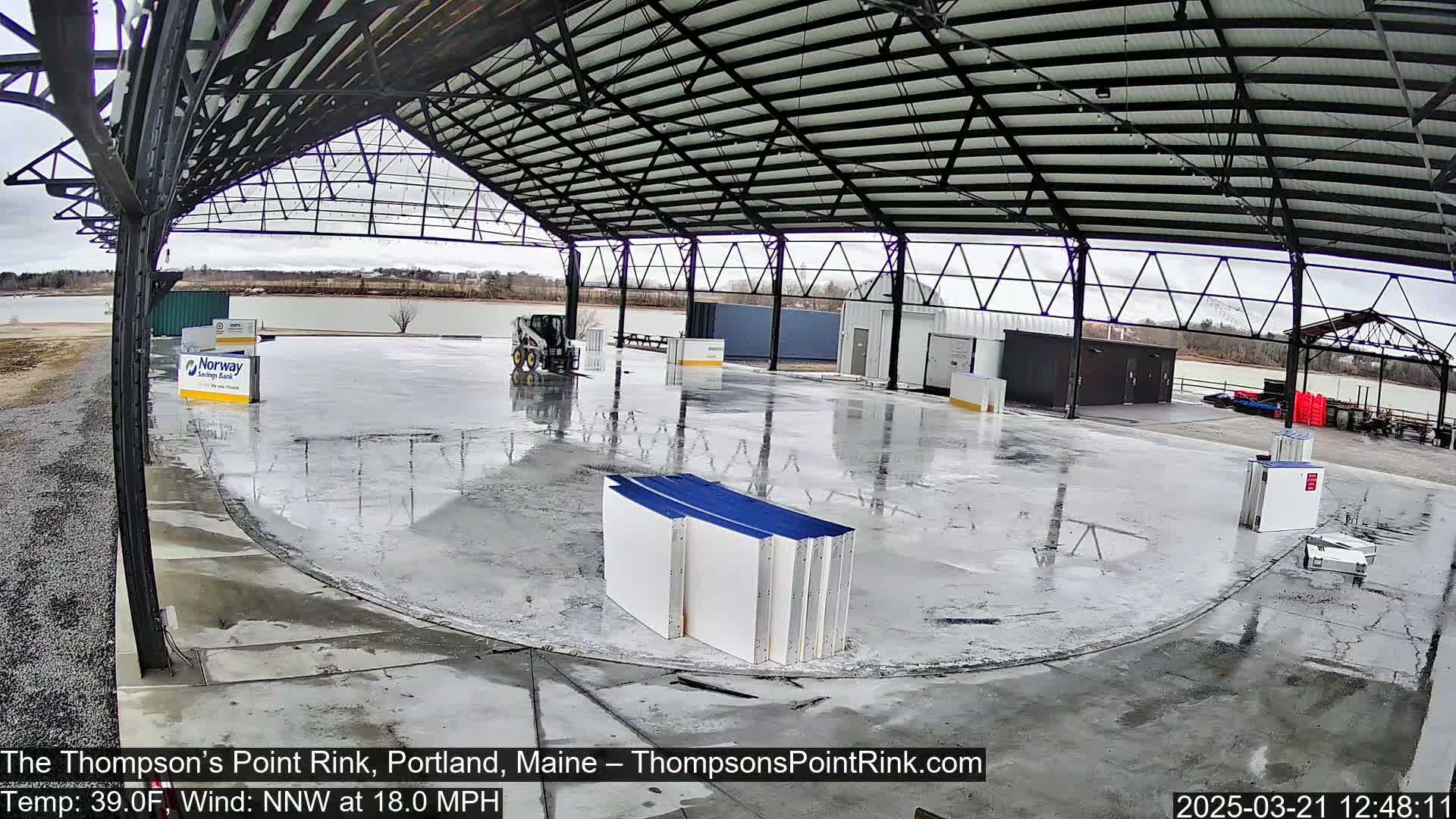 Under an overcast sky, a large, empty rink inside a metal structure is wet, with some building materials and a small piece of construction equipment present.