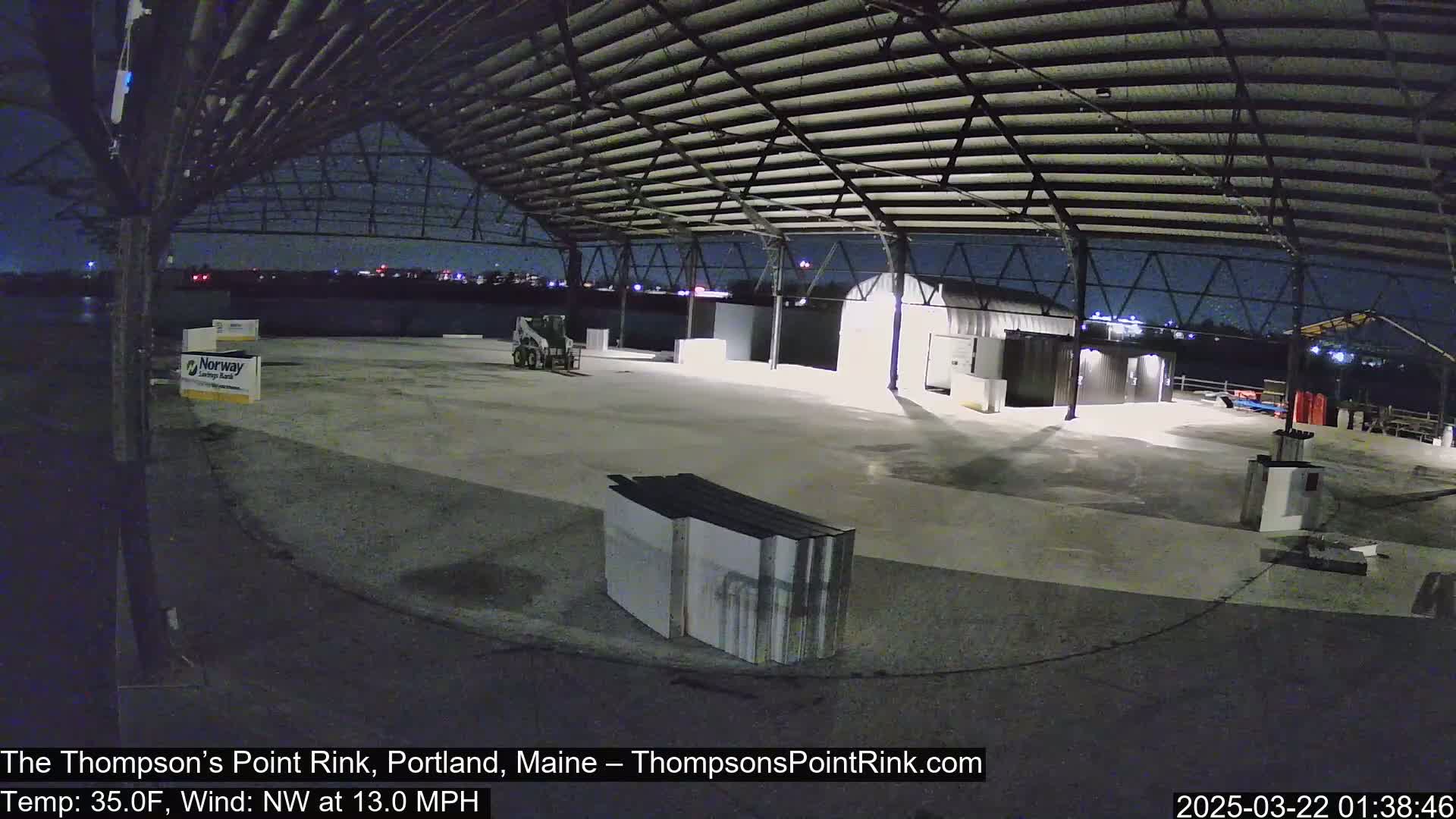 An empty, open-air rink under a metal roof at night, illuminated by artificial lights, with a northwest wind at 13 mph and a temperature of 35 degrees Fahrenheit.