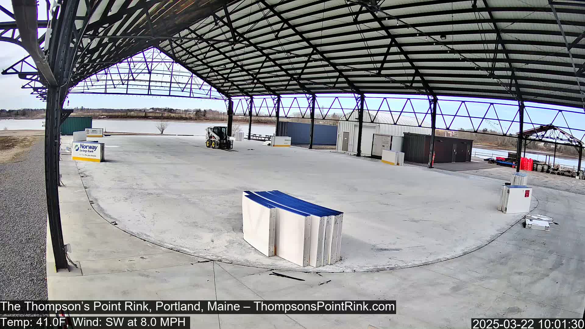 An empty paved rink under a metal-framed roof, with a partly cloudy sky and a body of water visible in the background.