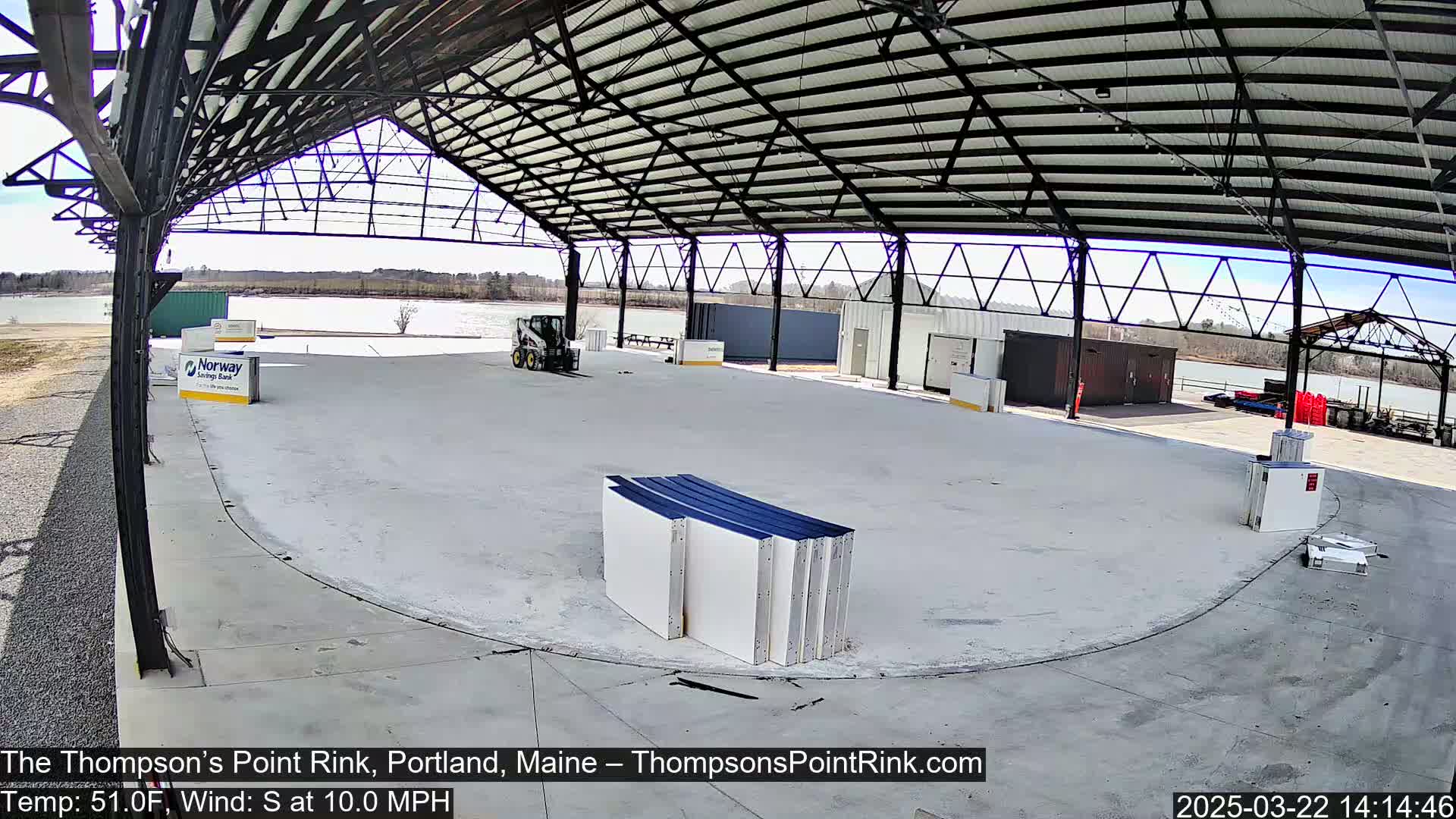 A large, empty indoor rink under a metal roof on a sunny day, with a body of water visible in the background.