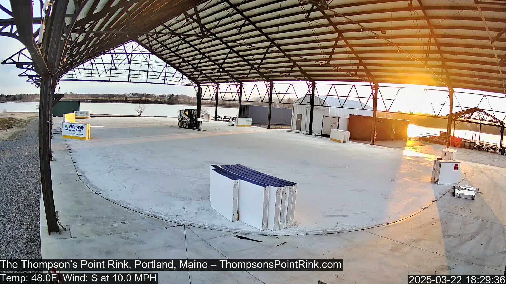 An empty rink under a large metal structure at sunset, with a calm body of water visible in the background.