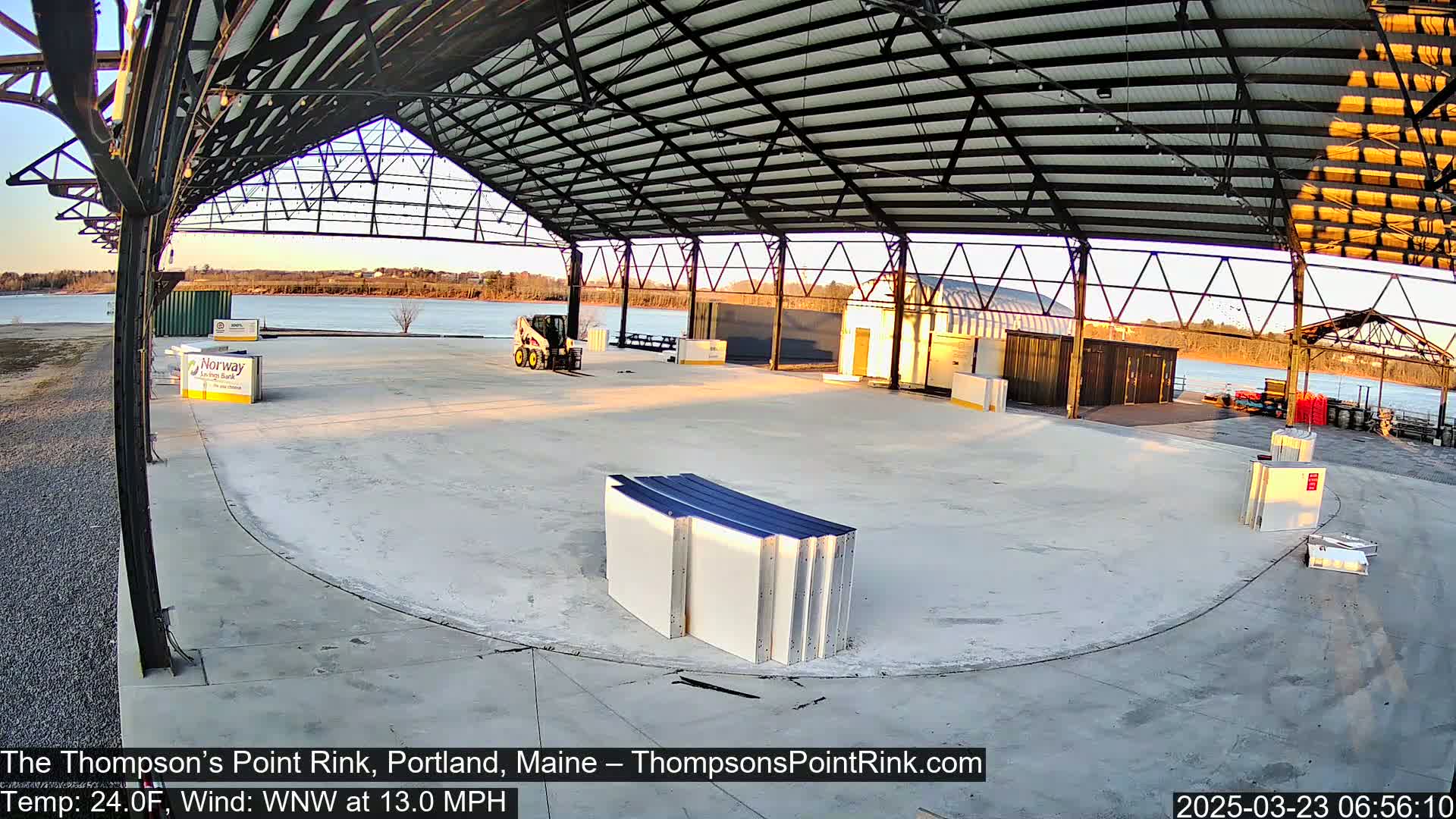 An empty, open-air rink under a metal roof, with a small piece of construction equipment and a calm body of water visible in the background at sunrise.