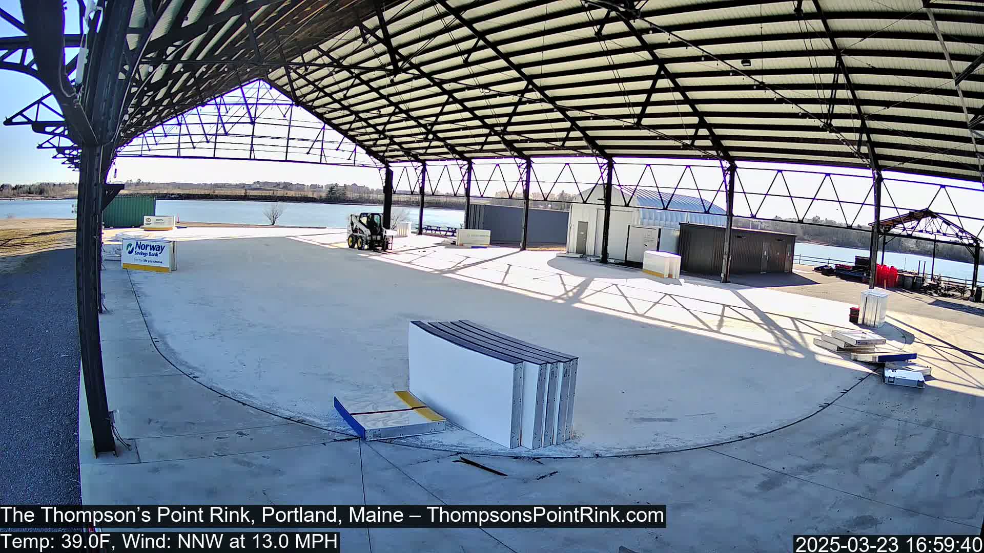 A partially constructed outdoor ice rink sits under a large metal roof on a sunny day with a 13 mph northwest wind and a temperature of 39°F.