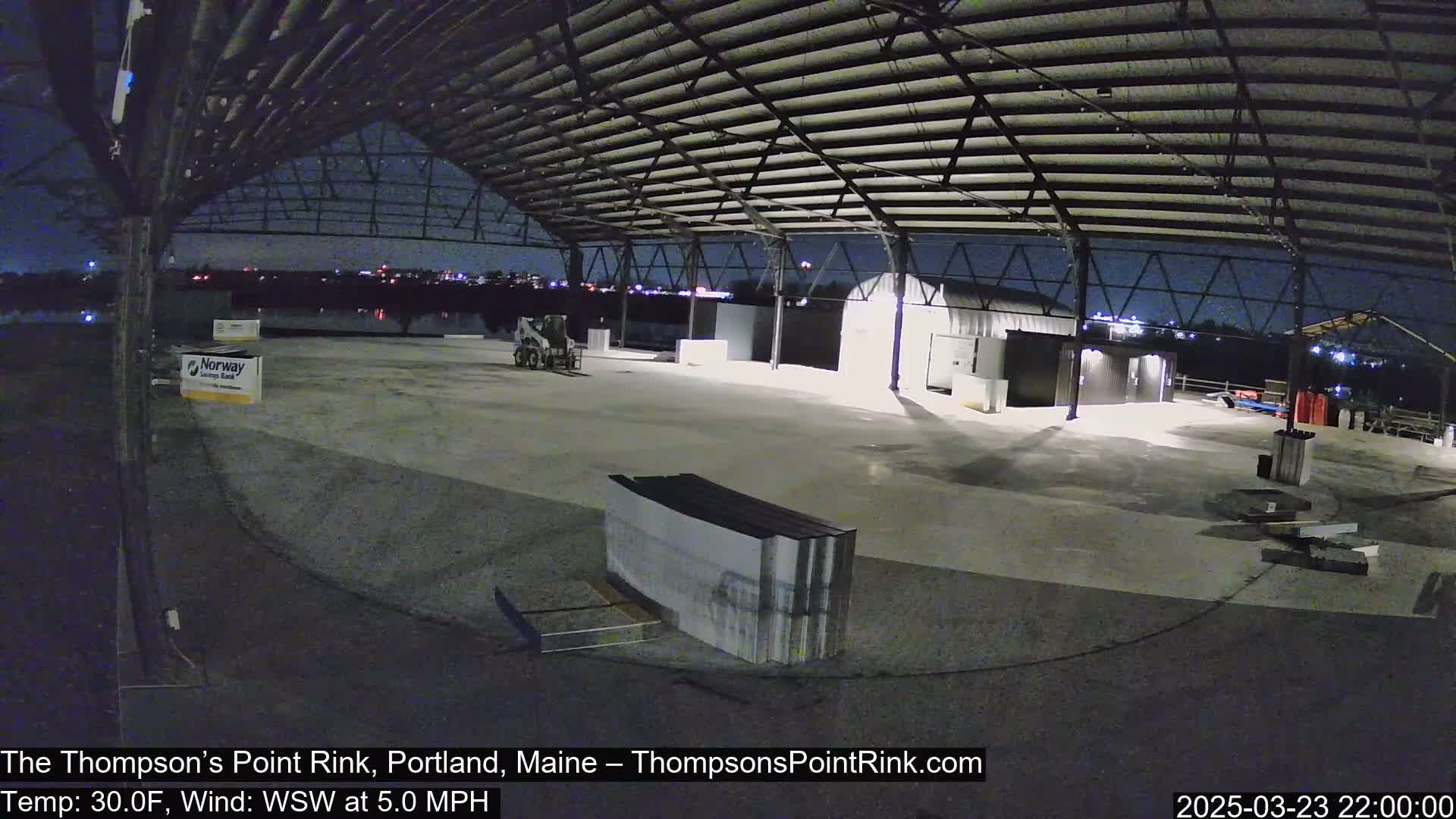 Under a clear night sky, a large empty rink sits under a metal roof and is surrounded by some small buildings and materials.