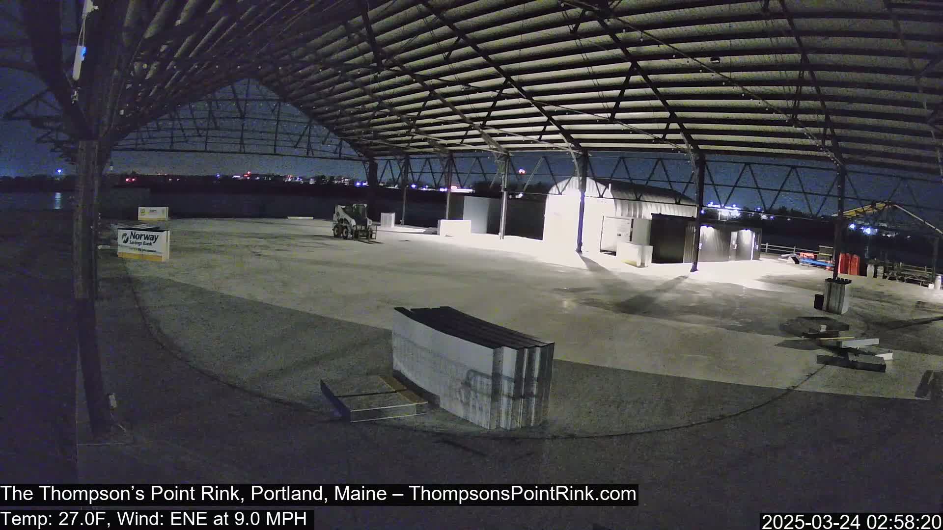 A large, empty, open-air rink under a metal roof at night, illuminated with some lights, under a clear, dark sky with a temperature of 27 degrees Fahrenheit and a 9 mph wind.