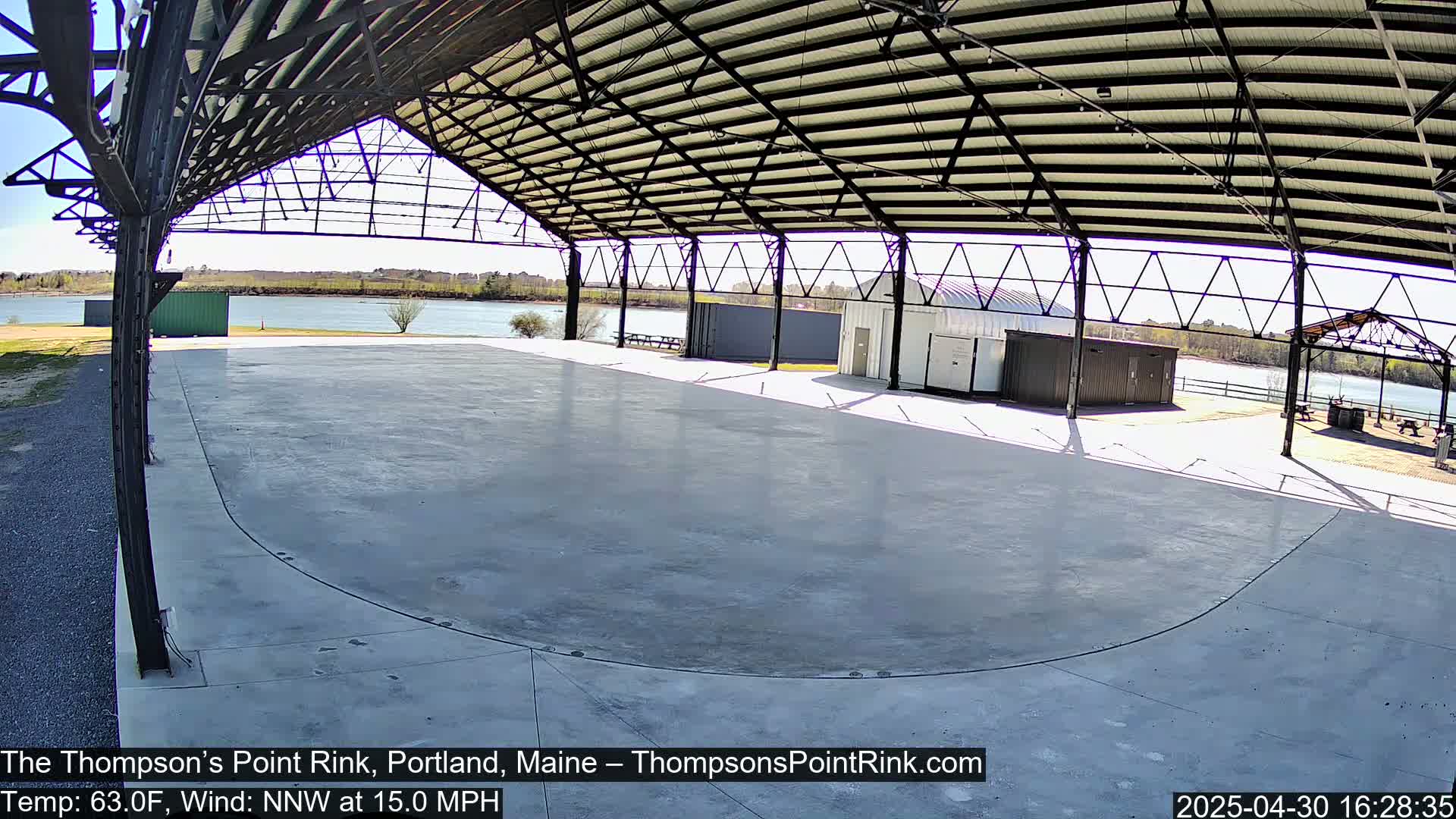 A large, empty, concrete rink sits under a metal-framed, open-air structure on a sunny day with a 15 mph northwest wind.