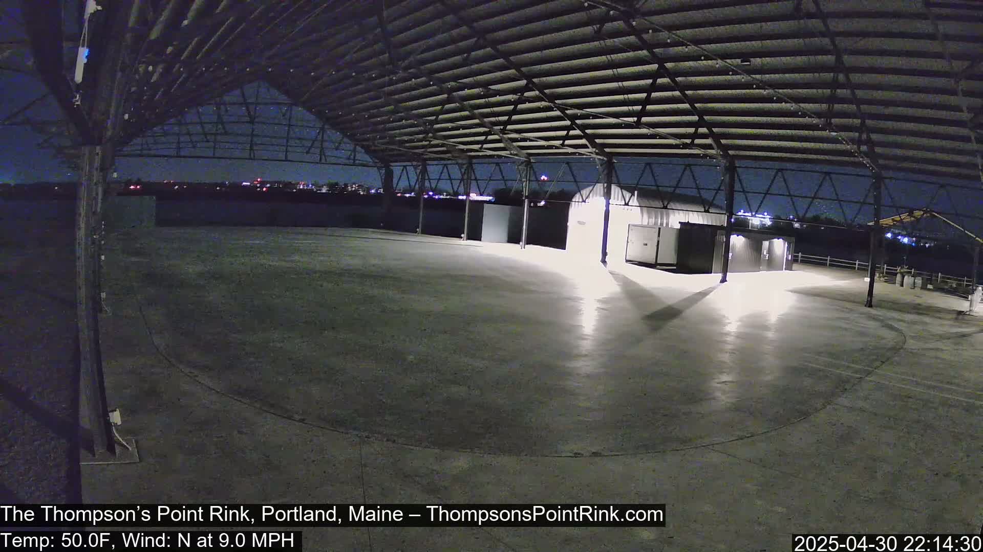 An empty, illuminated concrete rink sits under a metal roof at night, with a distant city skyline visible and a 9 mph north wind.