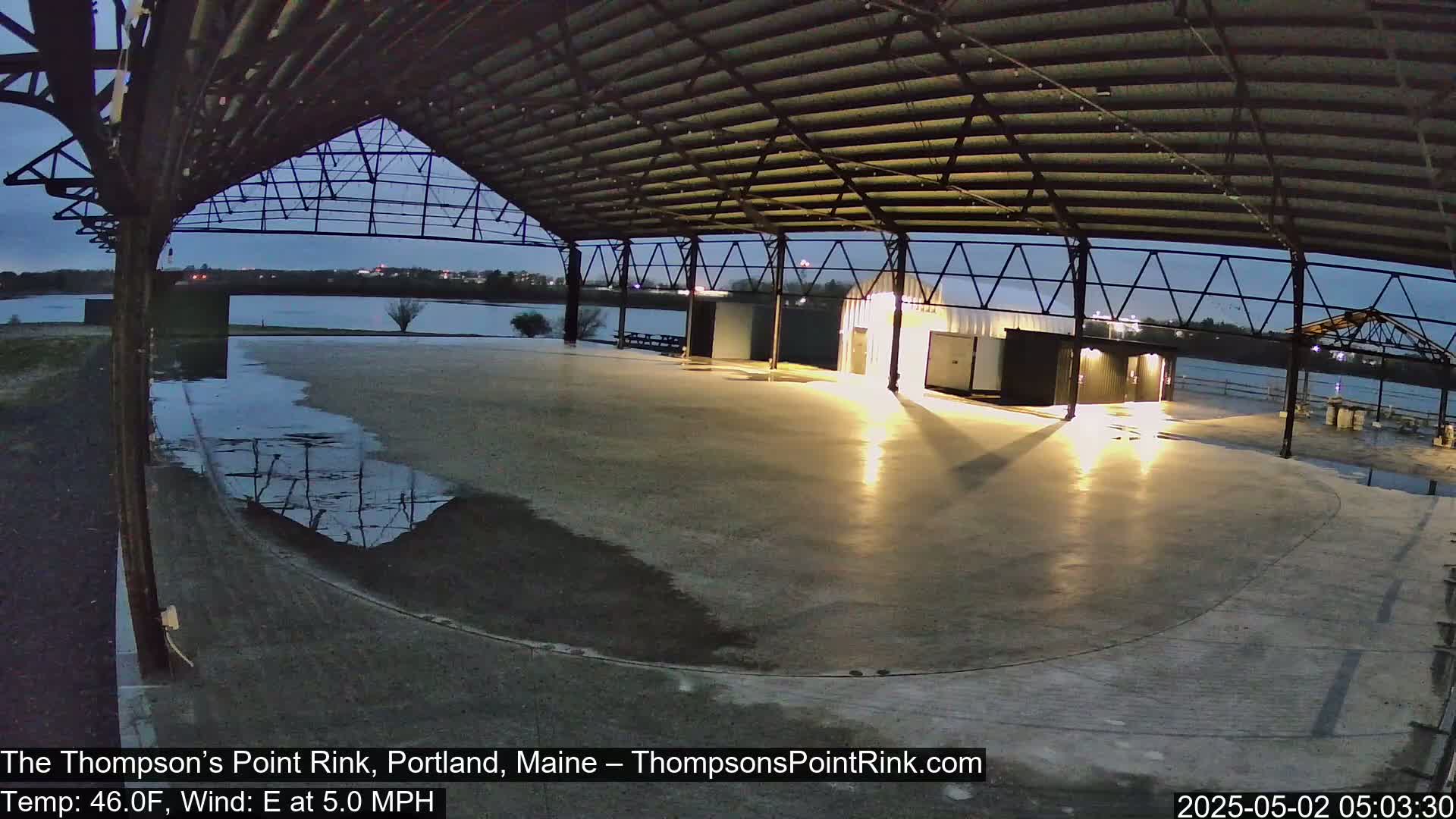 An empty, partially wet, concrete rink sits under a metal-framed structure at dusk, with a calm body of water visible in the background.