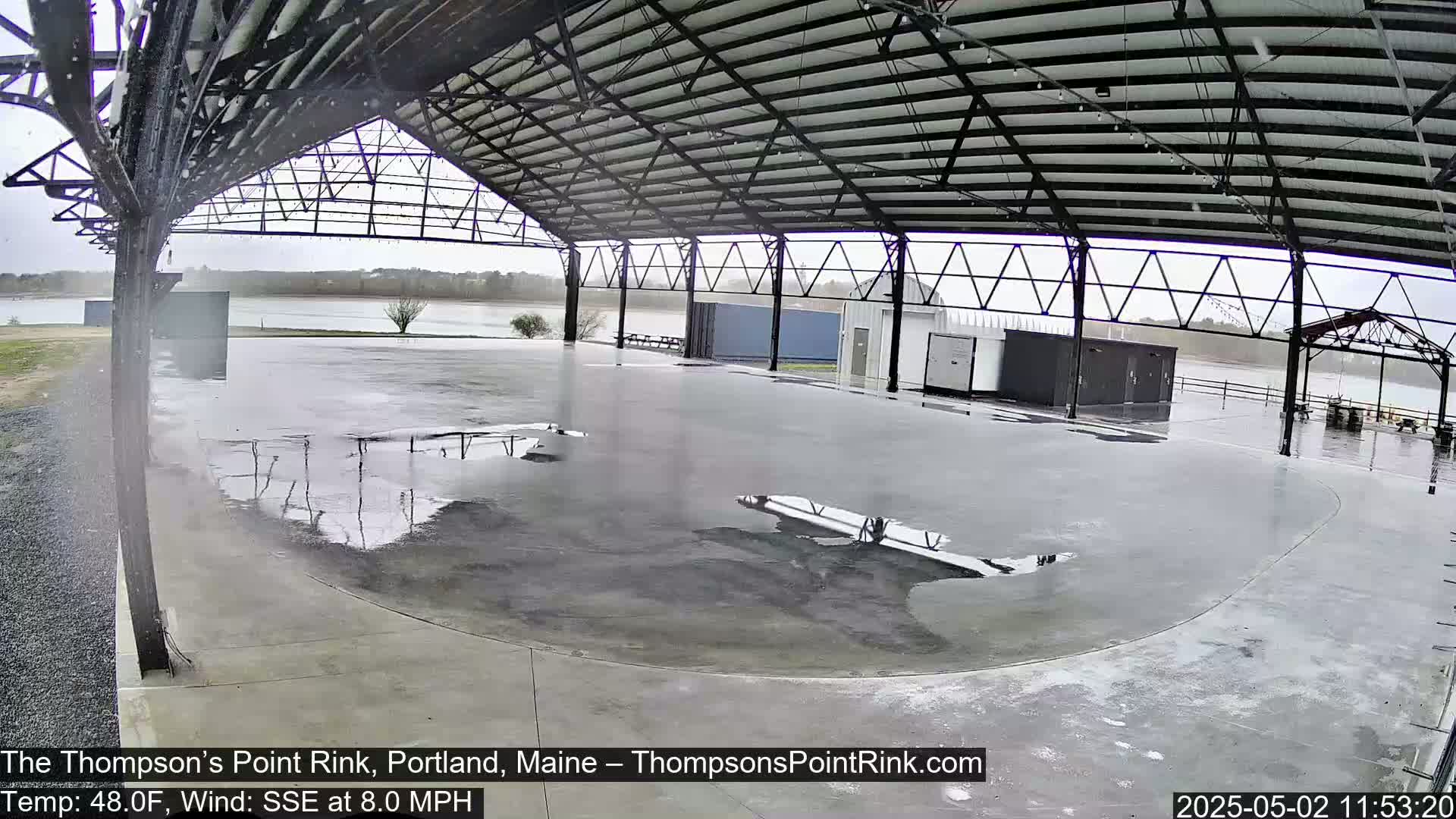 Rain falls on an empty, open-air concrete rink under a metal roof, overlooking a calm body of water.